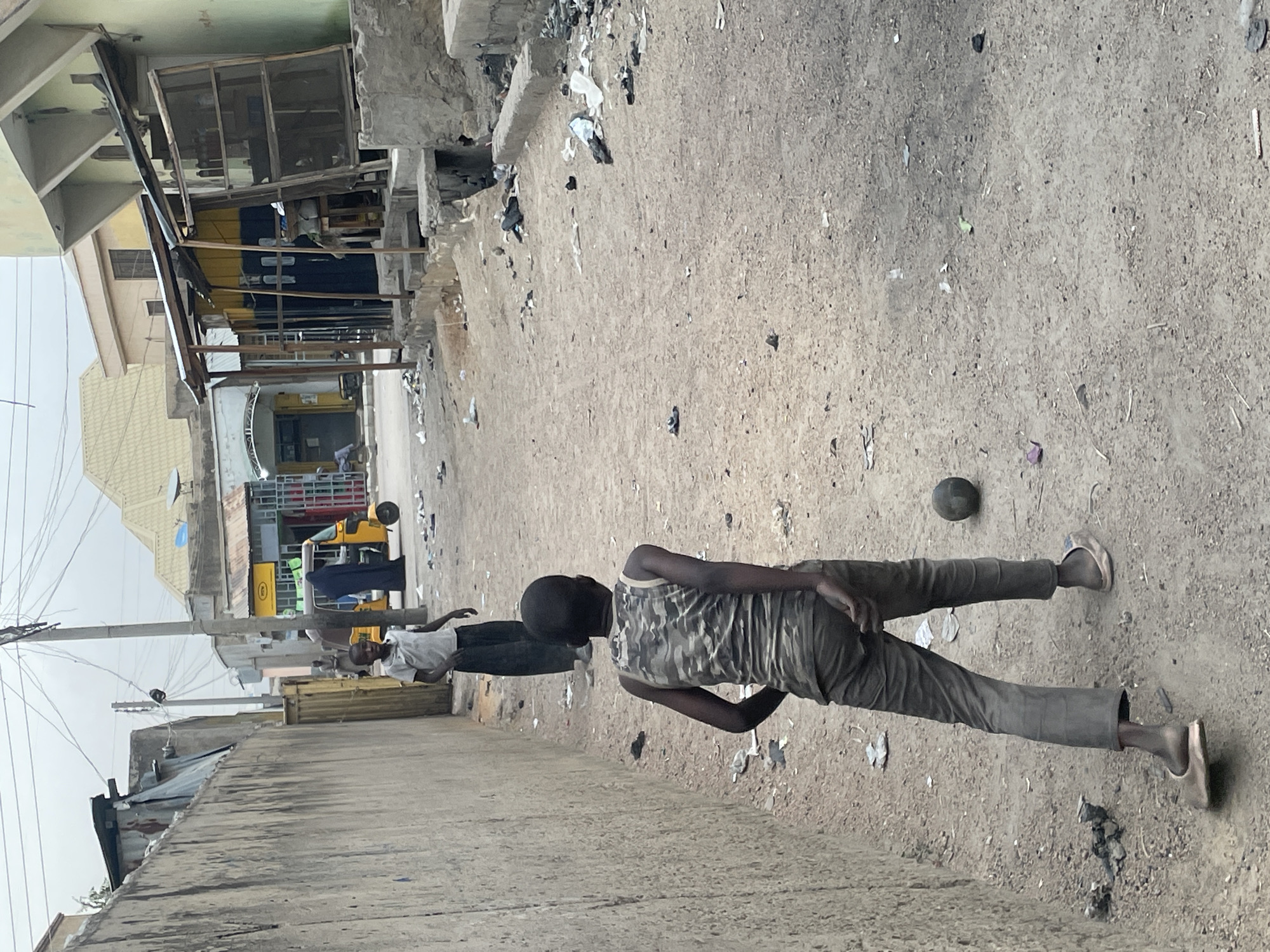 The image captures an outdoor scene on an unpaved, dusty street, likely during the daytime under an overcast sky. The ground is light brown dirt, heavily littered with various pieces of debris, including paper, plastic, and other refuse.
In the foreground, a person, presumably a man, is seen from behind, facing away from the camera. He wears a camouflage-patterned tank top and grey trousers, with light-colored slip-on shoes on his feet. His body is positioned as if in mid-stride or standing still, with his left foot slightly raised. Near his right foot, a dark, round object, possibly a ball or a stone, rests on the ground.
Further back and to the left, another person, also appearing to be a man, stands facing towards the left side of the frame. He wears a light-colored short-sleeved shirt and dark pants.
The background on the left side features several buildings of varying construction styles. Some have light-colored walls and metal roofs, while others show more weathered facades. Utility poles with numerous overhead wires crisscross the sky. A yellow and black tricycle, commonly known as a Keke Napep in some regions, is visible parked further down the street. The background on the right side shows a concrete or stone wall, partially crumbling, with exposed rebar, and more scattered debris along its base.
The overall setting suggests an urban or semi-urban environment, characterized by its unpaved roads, practical utility infrastructure, and visible signs of everyday life and neglect. There is no discernible text visible in the image.
