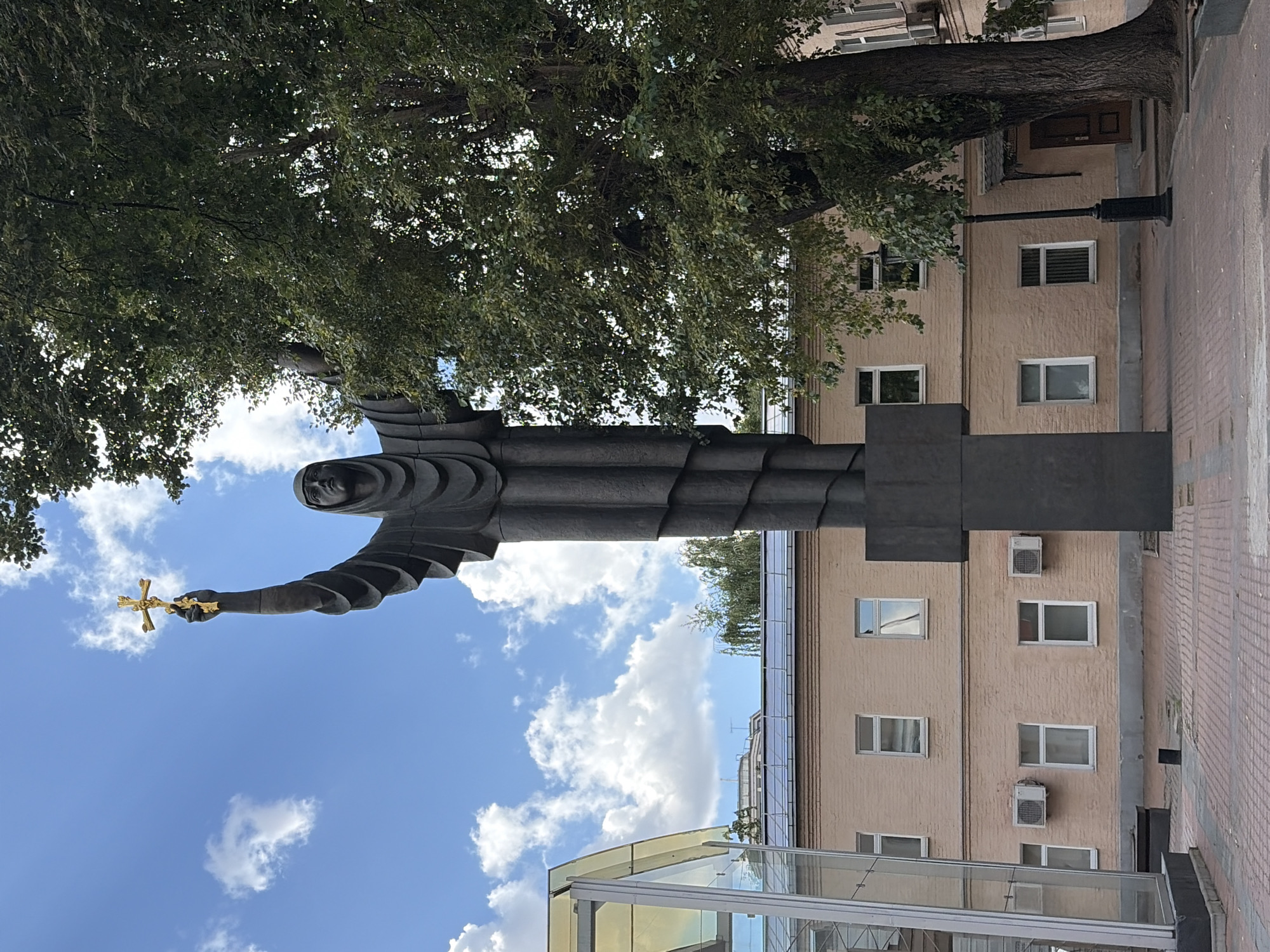 A monumental dark statue of a cloaked figure stands in the center of the frame, with its right arm raised and holding a golden cross. The statue is positioned in front of a pale brick building with multiple windows and air conditioning units. To the right of the statue, a large, leafy tree partially obscures the building. The sky above is blue with scattered white clouds. In the foreground, a patterned brick pathway leads towards the statue. A modern glass structure with a yellow accent is visible on the left side of the image, and a black lamppost stands near the tree. The location is the Patriarch's Ponds in Moscow, Russia.