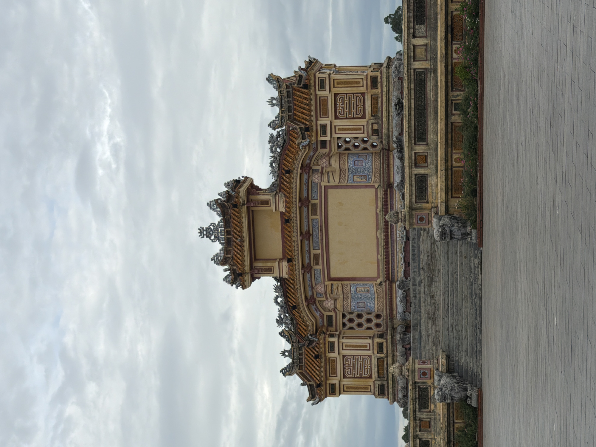 The image shows a frontal view of the Tomb of Khai Dinh, a mausoleum in the city of Huế, Thừa Thiên Huế Province, Vietnam. The structure is made of stone and wood, with a prominent central pavilion topped by a multi-tiered, ornate roof featuring dragon motifs. The facade is adorned with intricate carvings, decorative tiles, and yellow and red paint. A wide stone staircase leads up to the entrance, flanked by two large stone lions. The ground in front of the tomb is paved with large, grey stone tiles. In the background, the sky is overcast with grey clouds. Lush green foliage and some flowering plants are visible at the base of the structure. No people are present in the image.