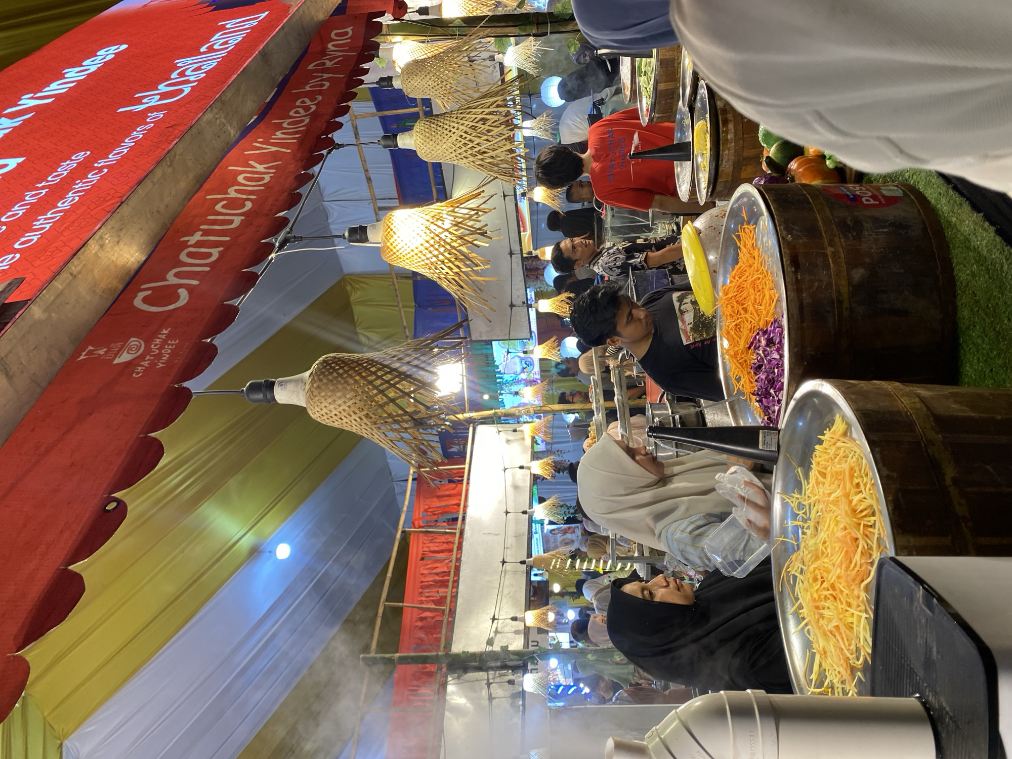 This image depicts a vibrant outdoor food stall, likely at a market, with a focus on fresh ingredients and food preparation. The scene is bustling with activity, with several people visible. In the foreground, a person wearing a black hijab is looking to the side, while another person in a white hijab is also visible. They appear to be engaged in some aspect of the food stall's operation, possibly serving or preparing food.
The stall itself is adorned with decorative bamboo lamps, casting a warm glow. Various food items are displayed, including shredded carrots and red cabbage in large metal bowls, suggesting a salad or stir-fry preparation. There are also whole vegetables like pumpkins and gourds visible. Wooden barrels are being used as display stands, adding to the rustic ambiance.
A prominent red banner with white text runs across the top of the image, reading "Chatuchak Yindee by Rina" and "e authentic flavors of". Another smaller banner below it, also red, has text and a logo, with the words "CHATUCHAK YINDEE" clearly visible. The setting appears to be a temporary outdoor structure, possibly a market or fair, indicated by the colorful fabric ceiling in yellow, white, and blue. The presence of mist or steam suggests cooking is actively happening. The overall mood is lively and engaging, typical of a busy food market. The specific location is identified as Lhokseumawe, Indonesia.