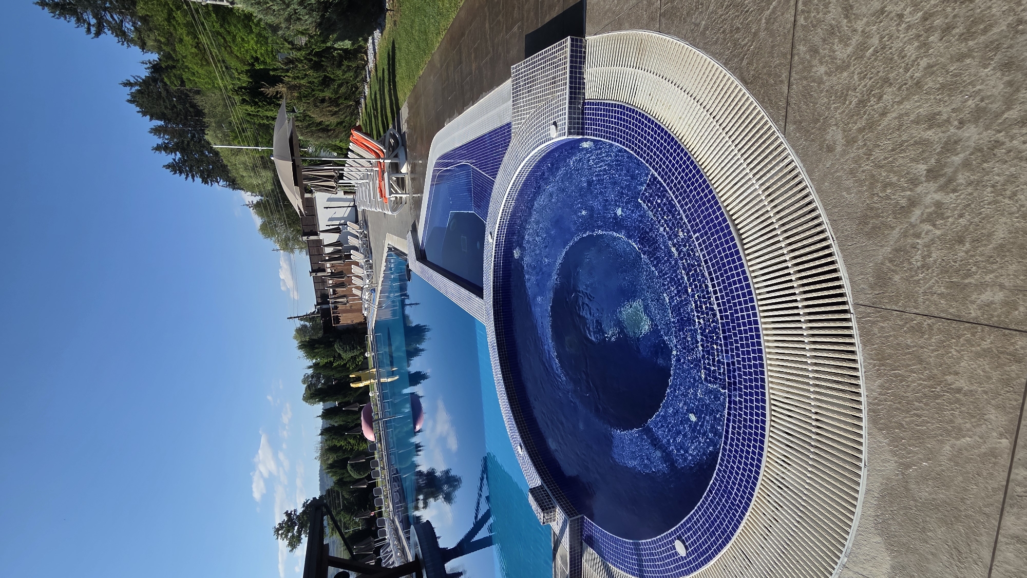 The image depicts an outdoor swimming pool and hot tub area on a clear, sunny day. The pool is large and rectangular with blue water. Adjacent to it is a smaller, circular hot tub filled with bubbling blue water, also tiled in blue. The surrounding area is paved with light-colored stone and features a white slatted drain system. Several lounge chairs and umbrellas are visible in the background, suggesting a resort or hotel setting. The scene is set against a backdrop of lush green trees and a blue sky. The location is identified as Bania, Bulgaria. There are no people visible in the image.