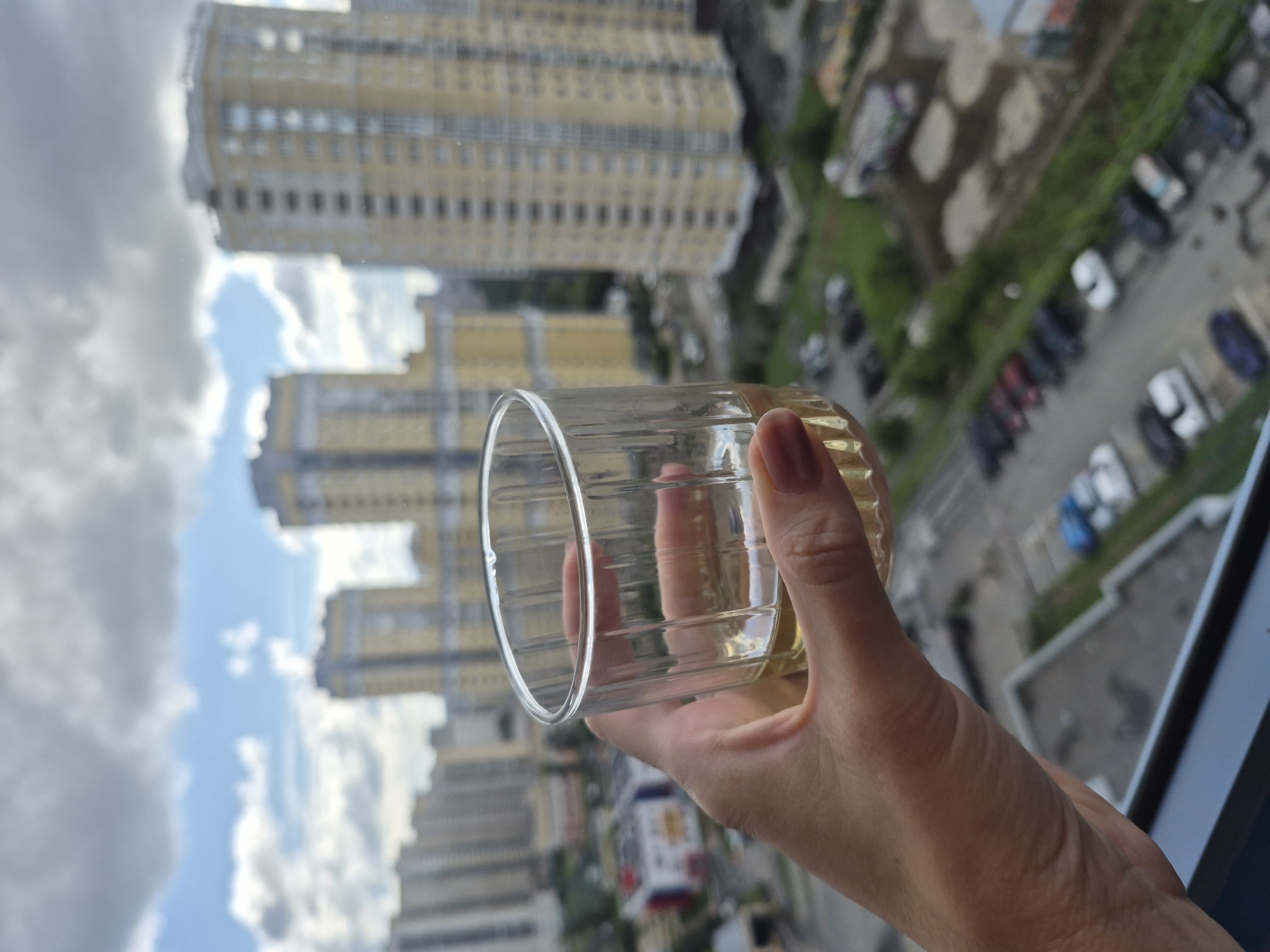 The image shows a person's hand holding a glass of amber liquid against a backdrop of a city skyline. The buildings are modern, tall, and appear to be residential high-rises, with a mix of yellow and grey facades. The sky is partly cloudy, with patches of blue visible. In the foreground, a parking lot with several cars can be seen. The perspective suggests the photo was taken from a high vantage point, likely an apartment balcony. The specific location within Yekaterinburg, Russia, cannot be determined from the visual information alone, as the architectural style and urban landscape are common to many large cities. The focus is on the hand and the glass, with the city serving as a blurred background.