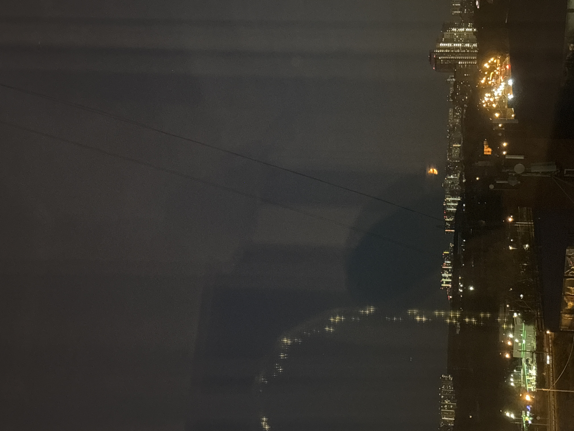 A nighttime, wide shot captures a cityscape with numerous lights from buildings and streetlights. The image appears to be taken from an elevated perspective, possibly a window, due to reflections visible on the surface. In the middle ground, there is a cluster of buildings with illuminated windows, suggesting residential or office spaces. Some of these buildings are taller, characteristic of urban environments. A highway or major road with moving vehicle lights is visible in the foreground, indicating traffic. There is a hint of a gas station with bright lights. The sky is dark and overcast. In the far distance, a faint, round, orange light is visible, possibly a distant building light or an atmospheric phenomenon. The overall scene depicts a typical urban night, with the glow of artificial lights illuminating the darkness. The location is the city of Odintsovo, Russia.