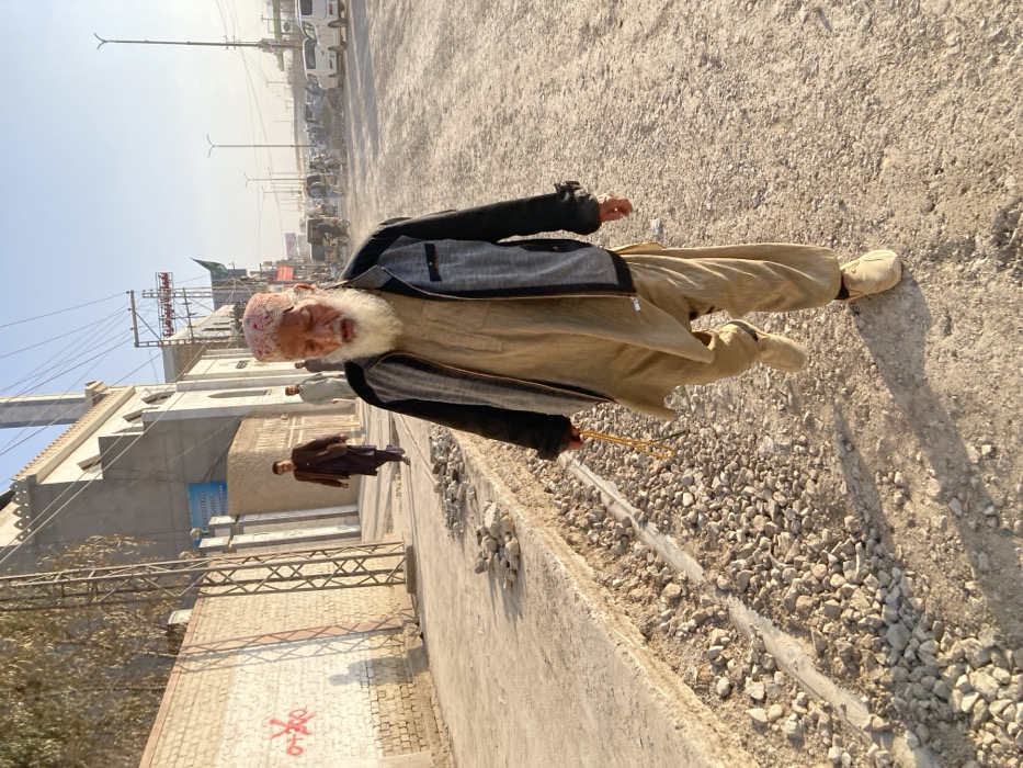 The image depicts an outdoor street scene in Quetta City Tehsil, Pakistan, under clear daylight conditions.
In the immediate foreground, an elderly male, appearing of South Asian ethnicity, walks towards the viewer on an unpaved surface composed of dirt and loose gravel. He wears a light tan traditional shalwar kameez, a grey and black zip-up jacket, and light-colored slip-on shoes. He has a prominent white beard and is wearing a red and white patterned skullcap. In his right hand, he holds a strand of yellow prayer beads. His gaze is directed downwards and slightly ahead.
To the left of the elderly man, an elevated concrete sidewalk runs parallel to his path. This sidewalk exhibits exposed layers of concrete and gravel, indicative of construction or erosion. Further left, a light-colored brick wall with red graffiti, including an 'X' symbol within a circle, is visible. A metal utility pole structure stands adjacent to this wall. Beyond the wall, a two-story building with light-colored walls and Islamic architectural features, including crenellated roofline elements and a visible minaret-like structure, appears to be a mosque. A blue sign is affixed to its exterior. Two additional males are observed walking on a paved section near this building in the midground, one wearing dark clothing and another in lighter attire further back.
The background on the right side of the image features a wide, dusty unpaved road extending into the distance. This road is occupied by numerous parked and moving vehicles, including multiple cars and what appear to be auto-rickshaws. Utility poles with overhead wiring and streetlights line this thoroughfare. The sky above is clear and bright, and direct sunlight casts visible shadows, suggesting a high sun angle.