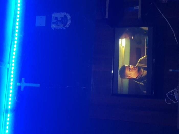 A television screen displays a scene with a man wearing a dark jacket. He's looking upwards with a focused expression. The room is dimly lit, with blue LED lights illuminating the left side. A cross hangs on the wall beside the lights. The overall atmosphere is dark and moody, with wires visible near the TV.