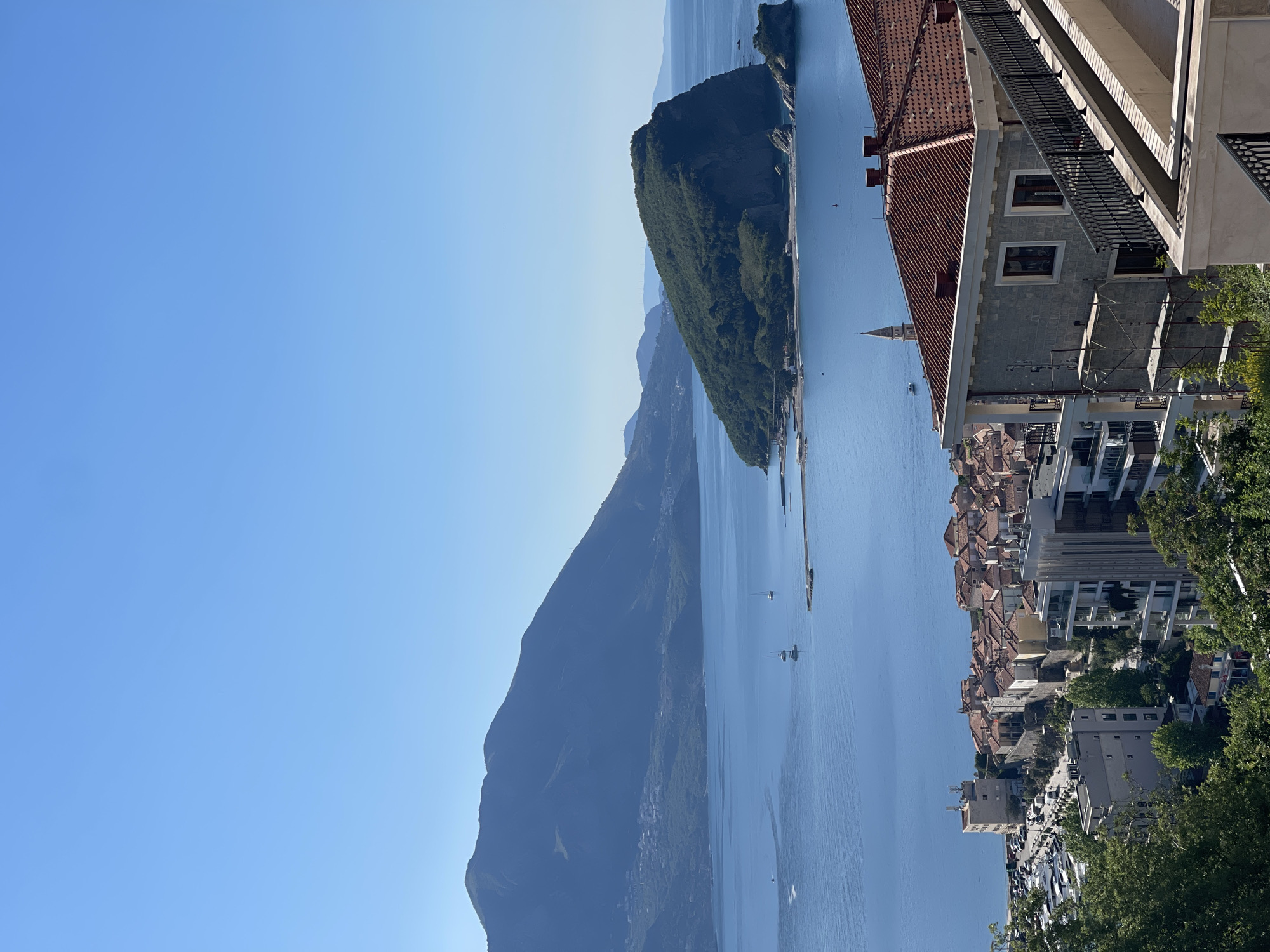 The image depicts a cityscape of Budva, Montenegro, with a view overlooking the Adriatic Sea. Mountains are seen across the bay in the distance. The foreground features a variety of buildings, including structures with red-tiled roofs and more modern constructions. A church spire rises amidst the buildings. Boats can be seen on the water. The sky is clear and blue. A building with a red tile roof and an adjacent building are located on the right side of the image.