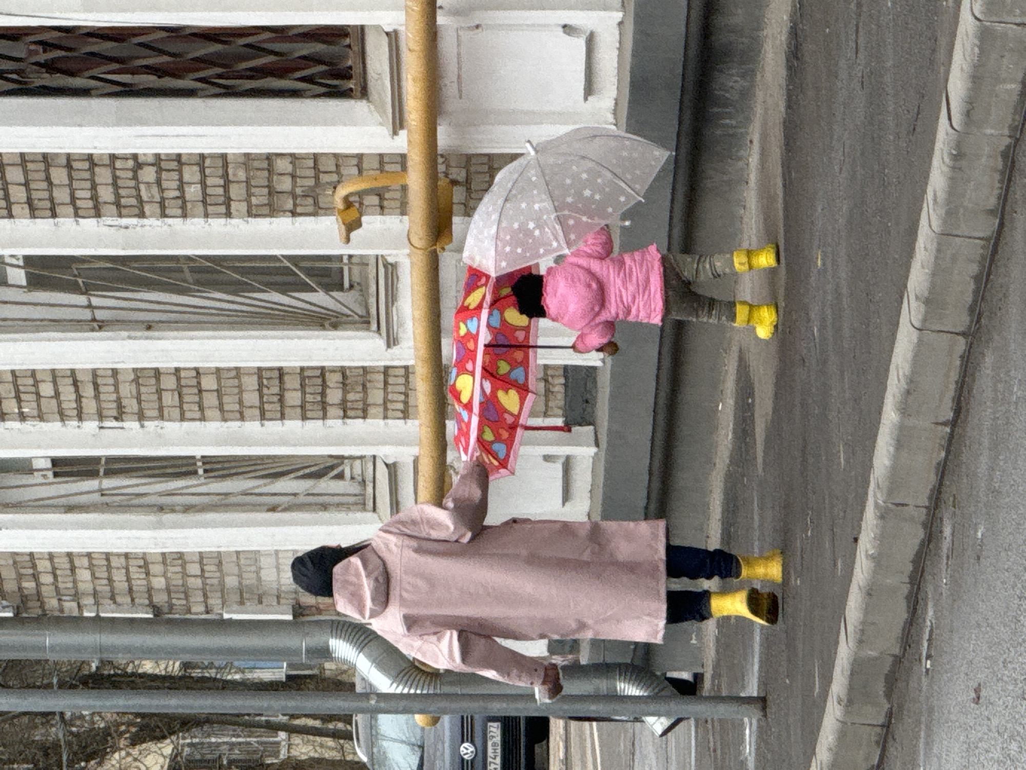 Two children wearing pink raincoats and yellow rain boots are walking on a sidewalk in Moscow, Russia. One child is holding a clear umbrella with stars on it, while the other is holding a red umbrella with hearts on it. The children are walking near a building with barred windows and a yellow gas pipe. A grey car is parked on the road.