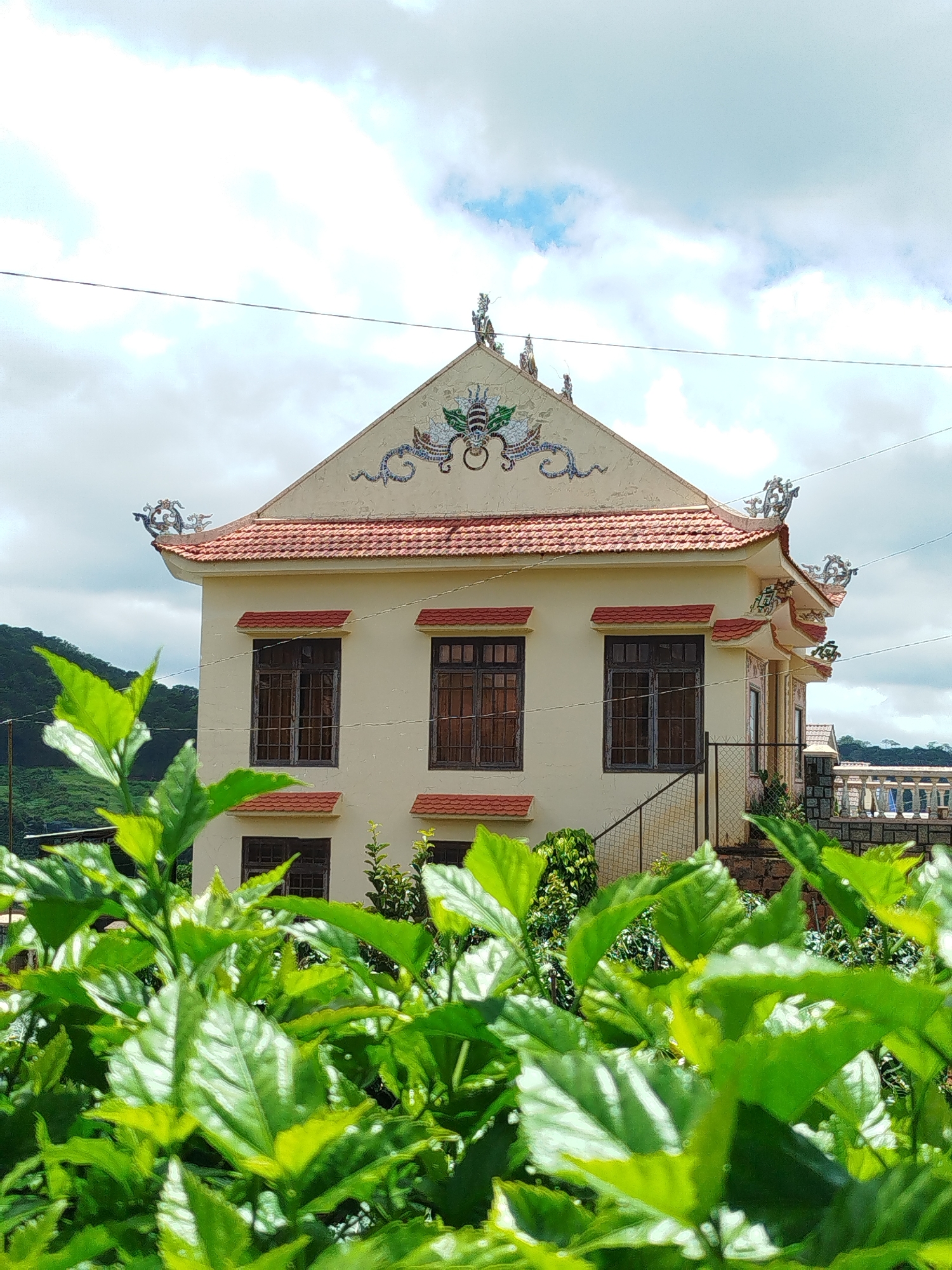 A traditional Vietnamese building, likely a temple or pagoda, stands against a cloudy sky. The building is painted a light yellow and features a red tiled roof with ornate dragon sculptures adorning the gables and eaves. There are several windows with dark wooden frames and red awnings. In the foreground, lush green foliage partially obscures the view of the building and its surroundings. The background shows a verdant hillside and a glimpse of another structure with a decorative balustrade. The scene is set in Xuân Sơn, Vietnam.