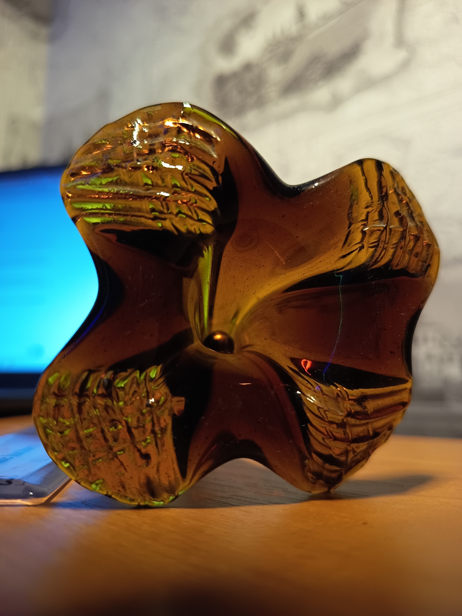 A close-up, low-angle shot captures a dark amber glass object with a star or flower-like shape. The object is positioned on a wooden surface, likely a table, with its textured surface catching the light, creating reflections of green and orange. In the background, a slightly out-of-focus blue light emanates from what appears to be a screen, possibly a computer monitor or television. Behind that, a textured wall with vertical lines can be seen. The lighting is warm and highlights the translucent quality of the glass. The image does not contain any information that can be used to determine its location in the city of Izmail, Ukraine.