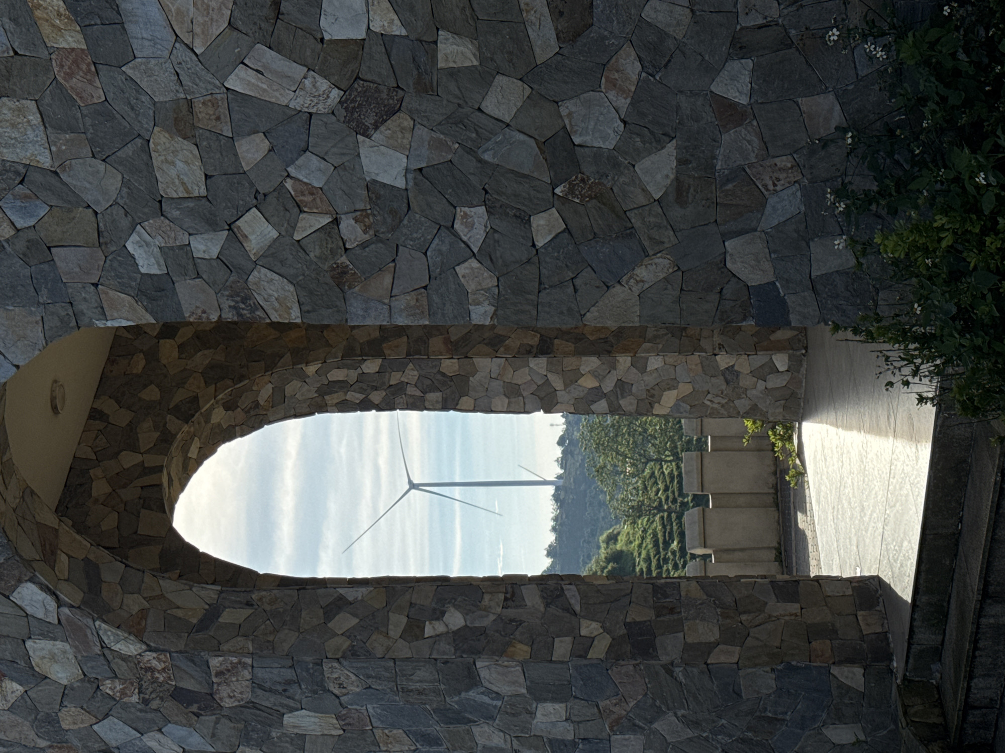 The image shows a stone archway with a view of a wind turbine in the distance. The archway is constructed from irregular, flat stones of varying shades of gray and brown. Through the arch, a landscape of rolling hills with terraced fields, likely tea plantations, is visible. A tall wind turbine stands prominently against a bright, partly cloudy sky. In the foreground, sunlight casts a bright reflection on the tiled ground, and some greenery with small white flowers is visible at the bottom right. The scene appears to be outdoors in a rural or semi-rural setting, possibly in the highlands of Phát Chi, Vietnam, known for its wind farms and agricultural landscapes.