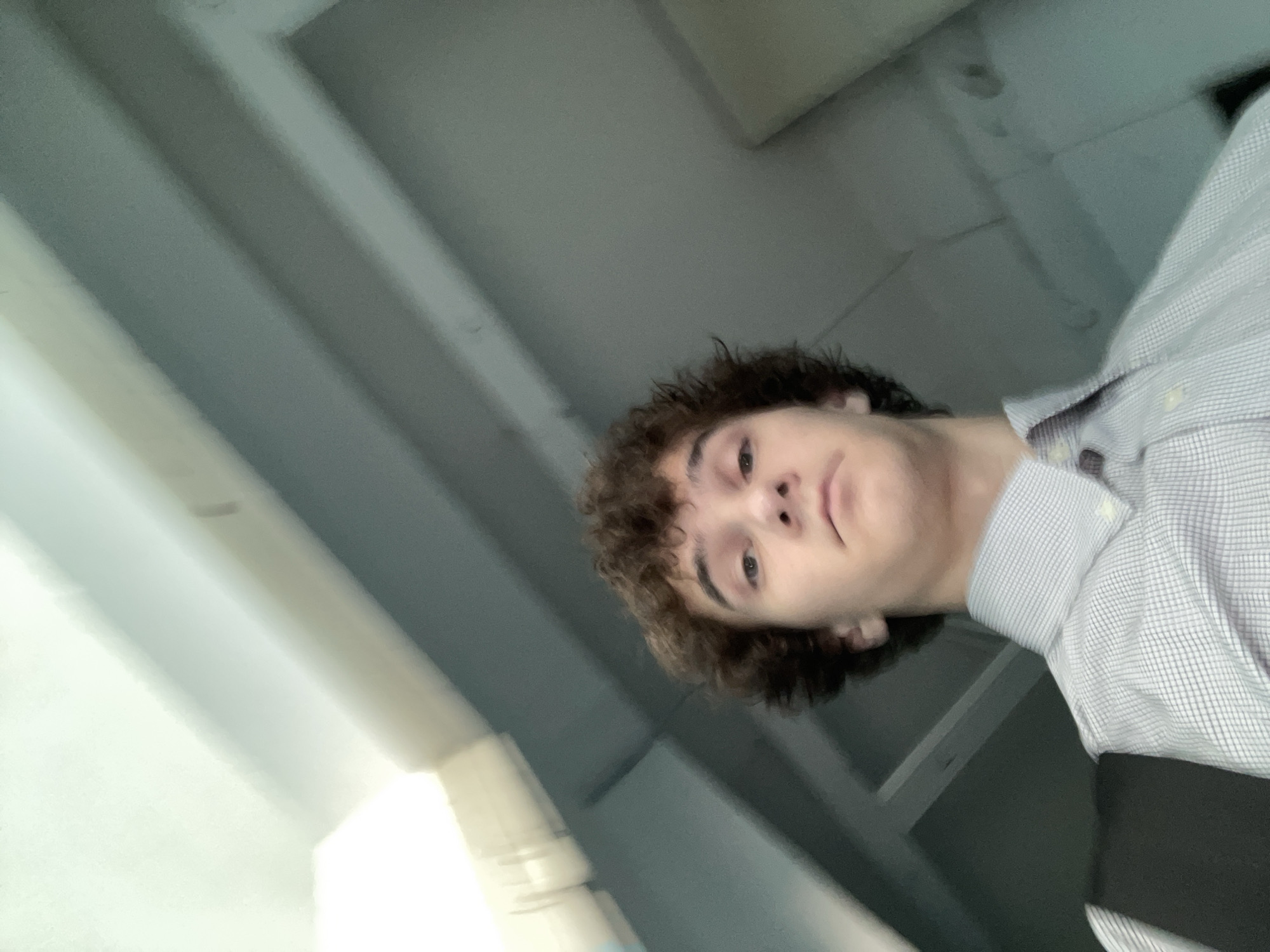 A low-angle, waist-up selfie of a young male with curly brown hair and light skin is shown. He is looking directly at the camera with a neutral expression. He is wearing a light blue and white checkered button-down shirt, partially obscured by a dark-colored strap across his chest, possibly from a backpack or bag. The background appears to be an indoor setting with a ceiling and some structural elements, possibly a bus or train interior, characterized by muted blue-grey tones and a bright light source at the top left. There is no indication of the location in Krasnoyarsk, Russia.