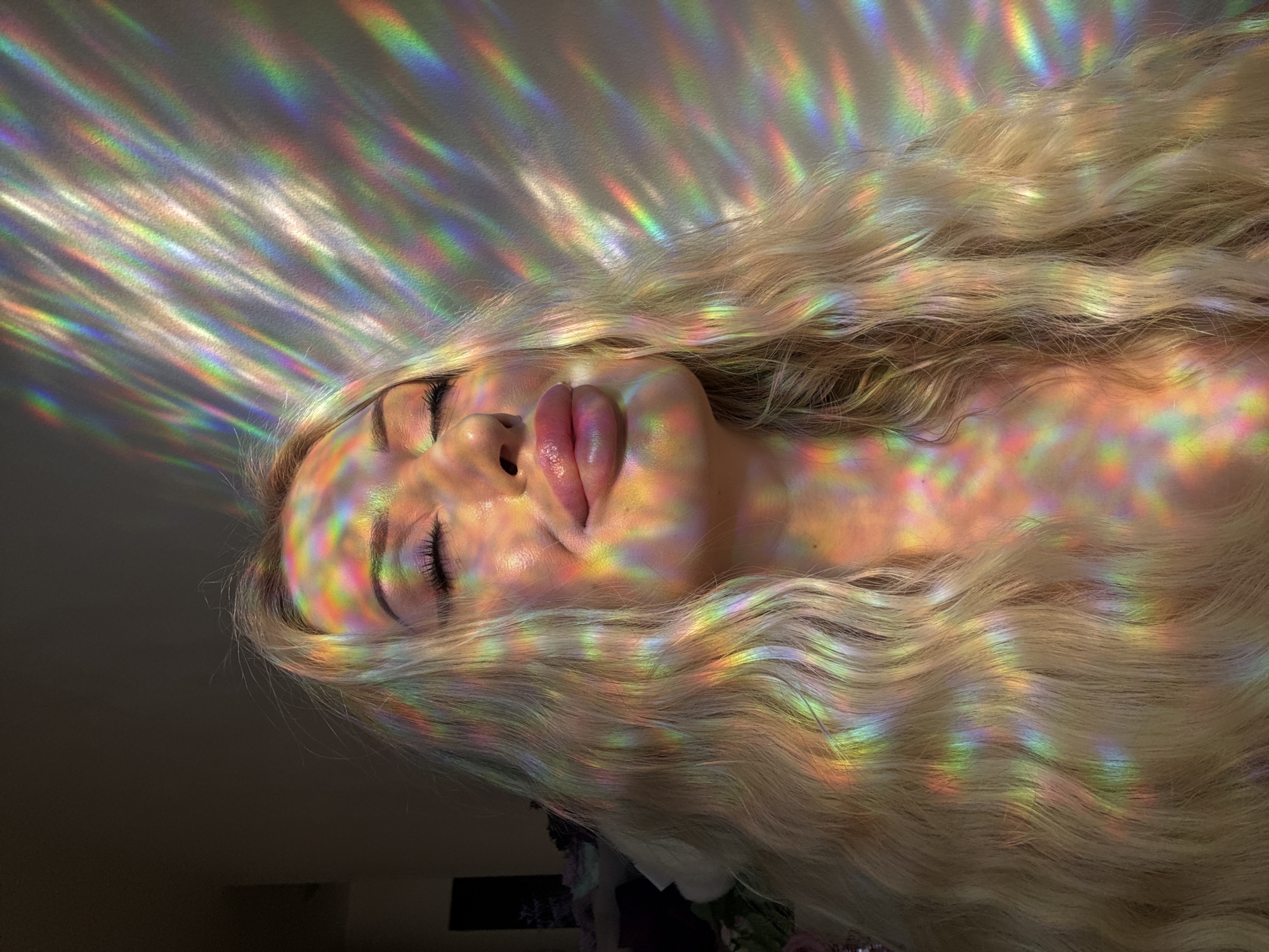 In a dimly lit room, a young woman with long, blonde, wavy hair poses with her eyes closed, her head tilted back. She is illuminated by a projected rainbow of light that casts colorful, striped patterns across her face, neck, and hair, as well as onto the wall behind her. Her lips are parted slightly, and she appears to be in a state of peaceful contemplation. The background is dark, with a hint of purple flowers visible on the left side of the frame. The overall impression is one of ethereal beauty and tranquility.