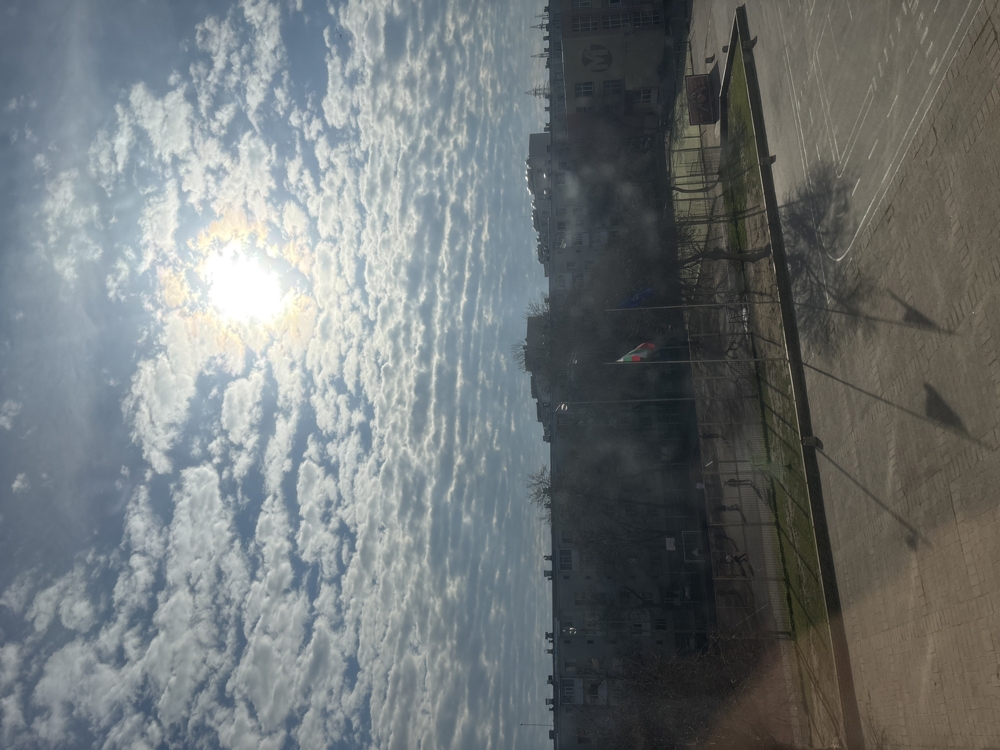 The image shows a cityscape in Plovdiv, Bulgaria, under a sky filled with altocumulus clouds and a bright sun. The foreground captures a schoolyard with painted markings on the pavement, adjacent to a fence enclosing a grassy area with trees. Buildings line the horizon, and flags are visible in the middle ground. The shadow of a flagpole is cast on the paved ground.