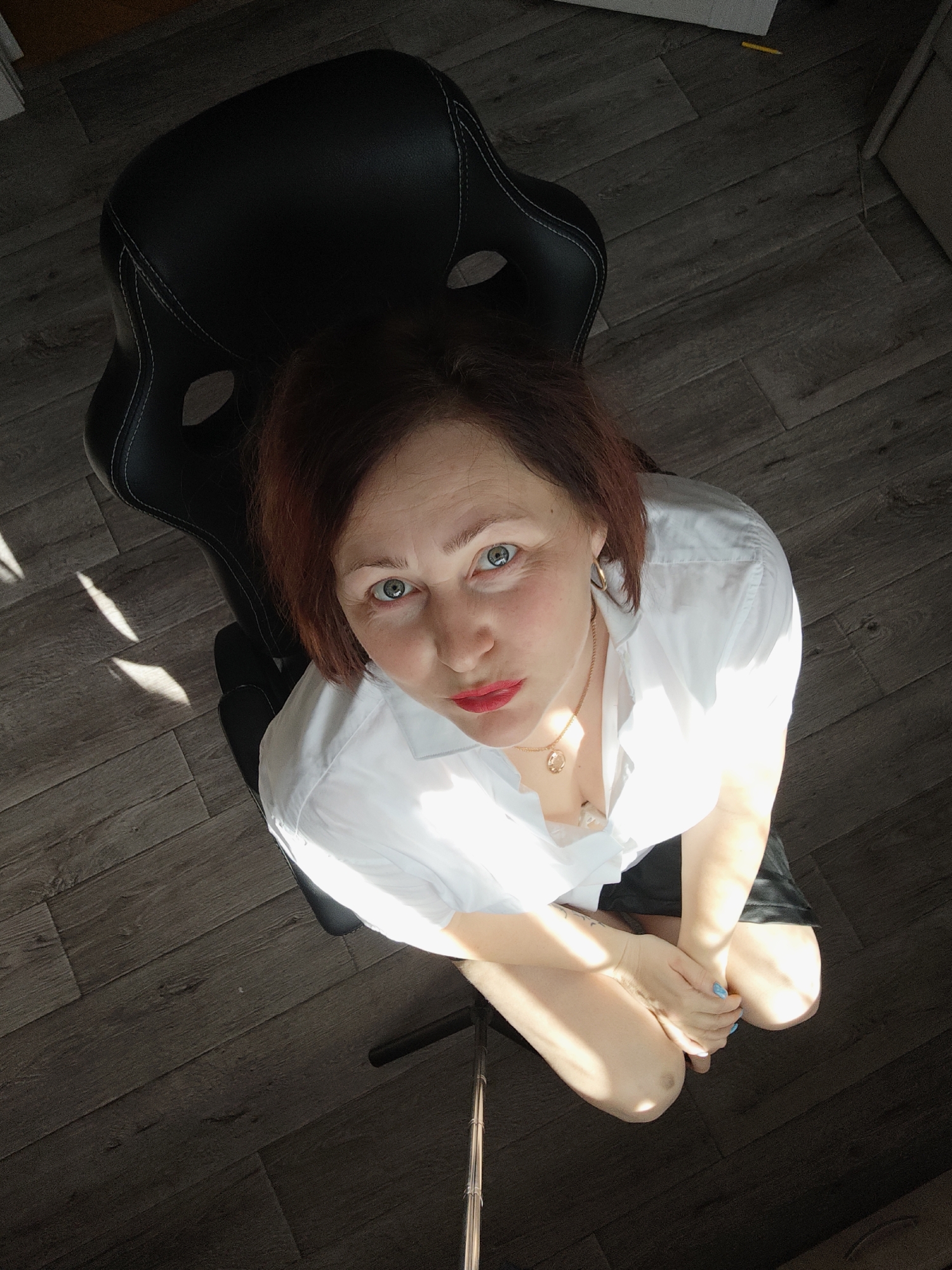 A high-angle shot shows a woman sitting in a black gaming chair, looking up at the camera. The woman has auburn hair, fair skin, blue eyes, and wears bright red lipstick. She is wearing a white collared shirt, and a gold necklace is visible at her collarbone. Her hands are folded in her lap, and her fingernails appear to be painted a light blue. The chair has white stitching details. The flooring is a dark, wood-look laminate. Sunlight streams across the scene from an unseen source, casting shadows on the floor and the woman. The photo appears to be taken indoors, possibly in an office or home setting in Марковский, Russia.