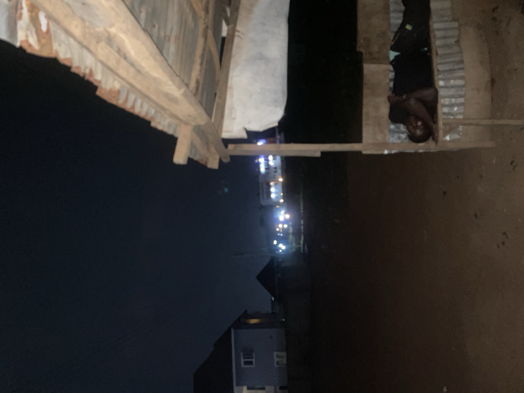 The image depicts a nighttime scene in what appears to be a low-income area of Abuja, Nigeria. The view is partially obscured by a rough wooden structure, possibly part of a makeshift shelter or unfinished building. A person is lying down near the wooden structure, and a building and streetlights are visible in the distance. The lighting is poor, suggesting the area is not well-lit.