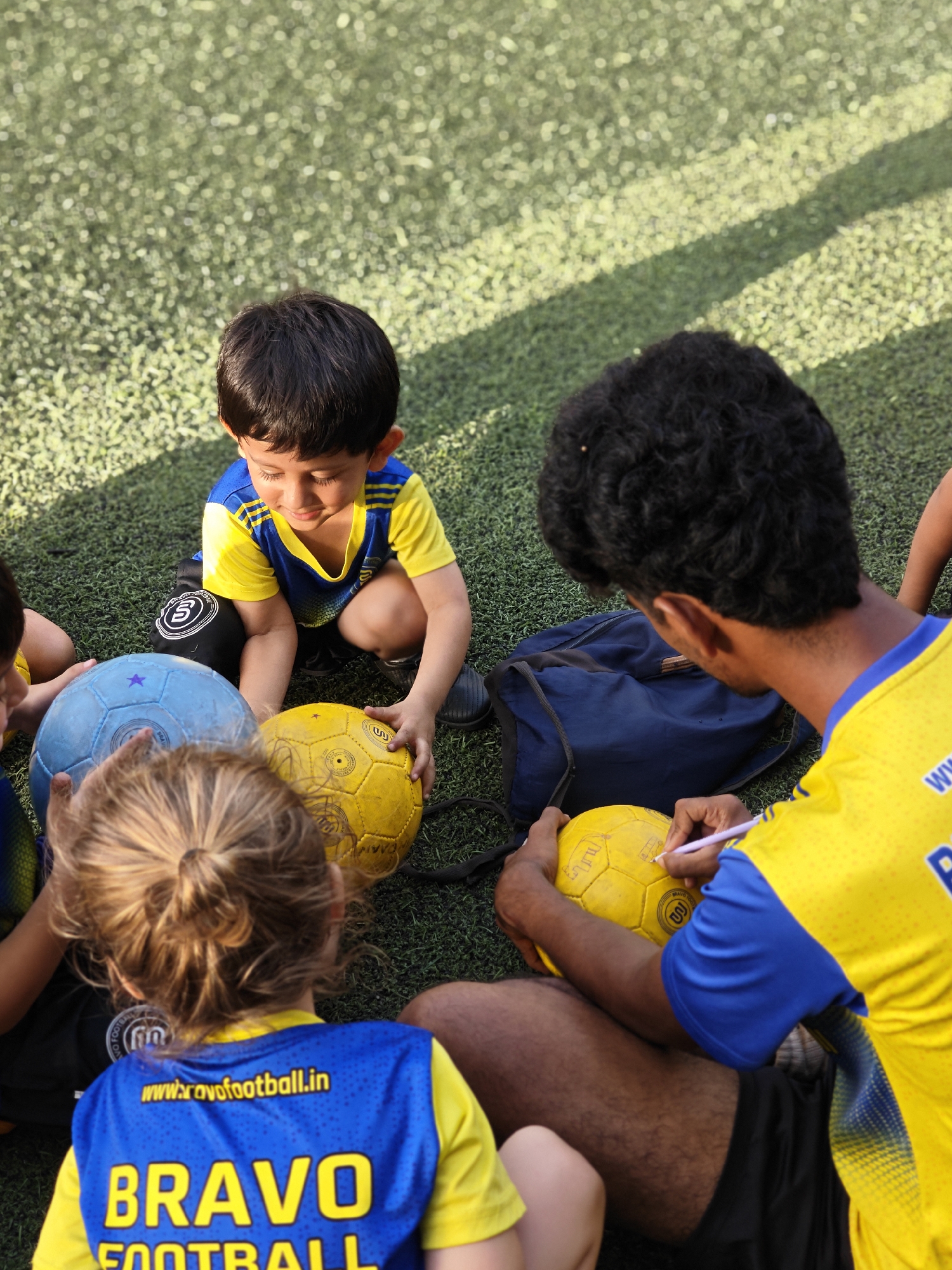 A medium shot captures a group of individuals on an artificial turf field. A young child in a blue and yellow sports jersey squats, holding a yellow soccer ball. Another child with light hair and a similar jersey is partially visible in the foreground. An adult male, also wearing a matching blue and yellow jersey, sits on the ground, writing on a yellow soccer ball with a pen. A blue backpack sits nearby on the grass. The back of the child's jersey in the foreground reads "BRAVO FOOTBALL," and the website "www.bravofootball.in" is printed above. Based on the website domain ending in ".in", it is likely the photograph was taken in India. Given the details on the jersey, it is probably a location within Mumbai, India.