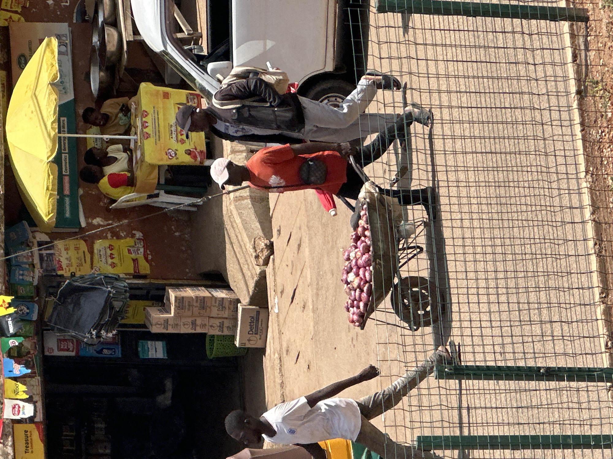 The image depicts a bustling street scene in Bukuru, Nigeria. In the foreground, a young boy in a white t-shirt walks beside a wheelbarrow filled with red onions. He is on the left side of the frame, his body in profile, and he appears to be walking away from the viewer. Behind him, two men are walking in the same direction. The man closer to the wheelbarrow wears an orange t-shirt and a dark crossbody bag, while the man to his right is in a dark jacket and light-colored pants, carrying a backpack. A white vehicle is parked on the right side of the image, partially obscuring the background.
In the middle ground, a market stall is set up with a yellow umbrella providing shade for a table where three people are seated. The stall has various items displayed, and signs are visible on the wall behind it. The ground appears to be a mixture of dirt and pavement. The overall impression is that of a busy, commercial area with people engaged in trade and movement.