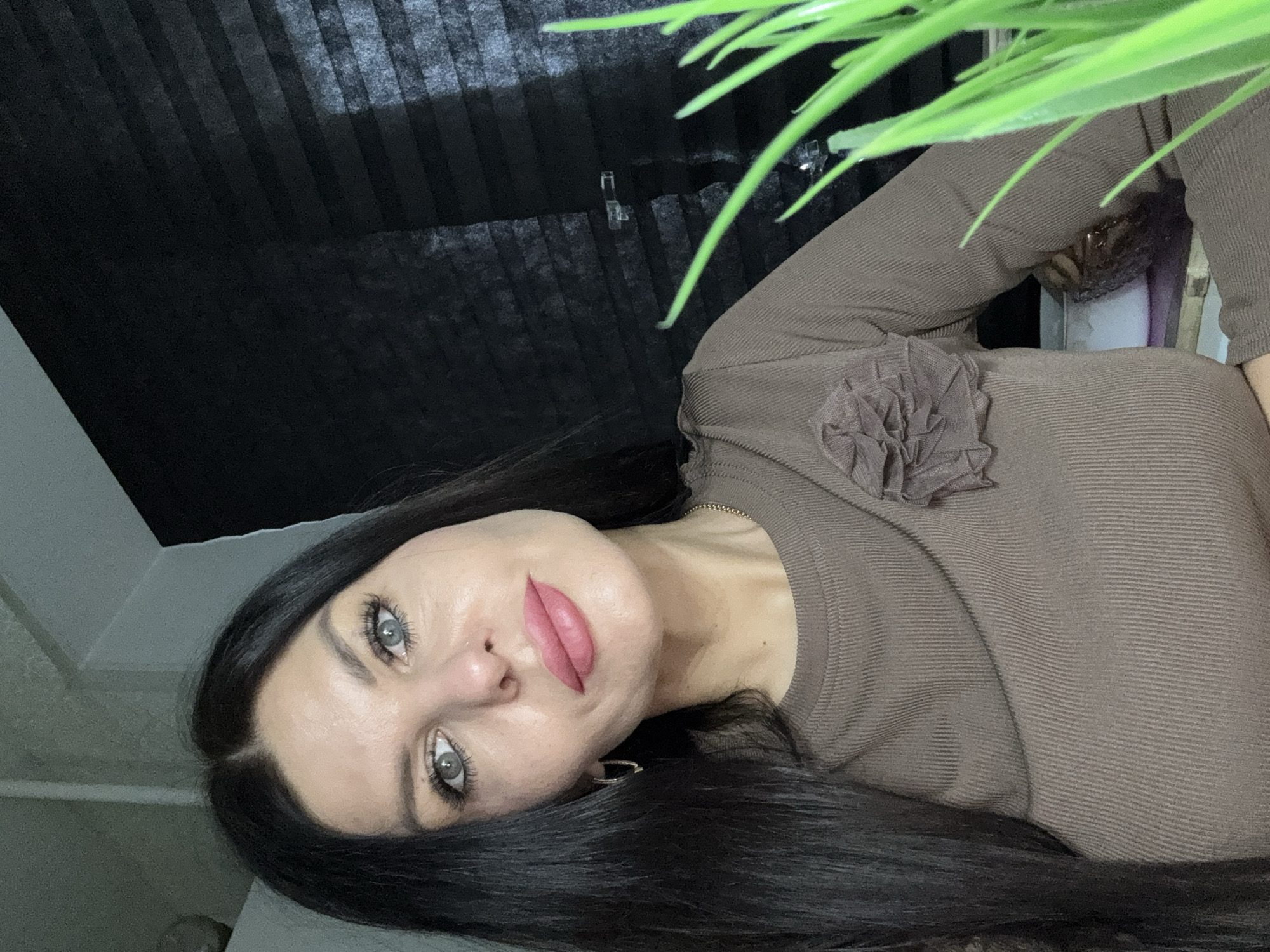 A woman with dark hair and light eyes is facing the camera. She has full, light pink lips and is wearing a brown, ribbed top with a ruffled flower detail on the upper chest. A partial view of green plant leaves is visible at the top right. The background features a dark, textured surface, possibly window blinds. The setting and location information, specifically Chaikovsky, Russia, are not discernible from the image alone.