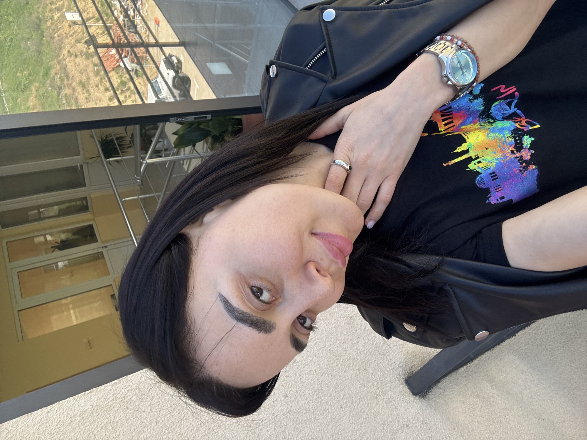 A woman with dark hair and light skin is angled in the foreground, wearing a black t-shirt with a colorful, cityscape-inspired graphic and a black leather jacket. She has a silver ring on her finger and a silver watch with a beaded bracelet on her wrist. Behind her is a building with glass windows and a railing, as well as a partial view of a green and brown landscape. The scene appears to be located on a balcony or outdoor space in Tbilisi, Georgia, based on the architectural style of the building.