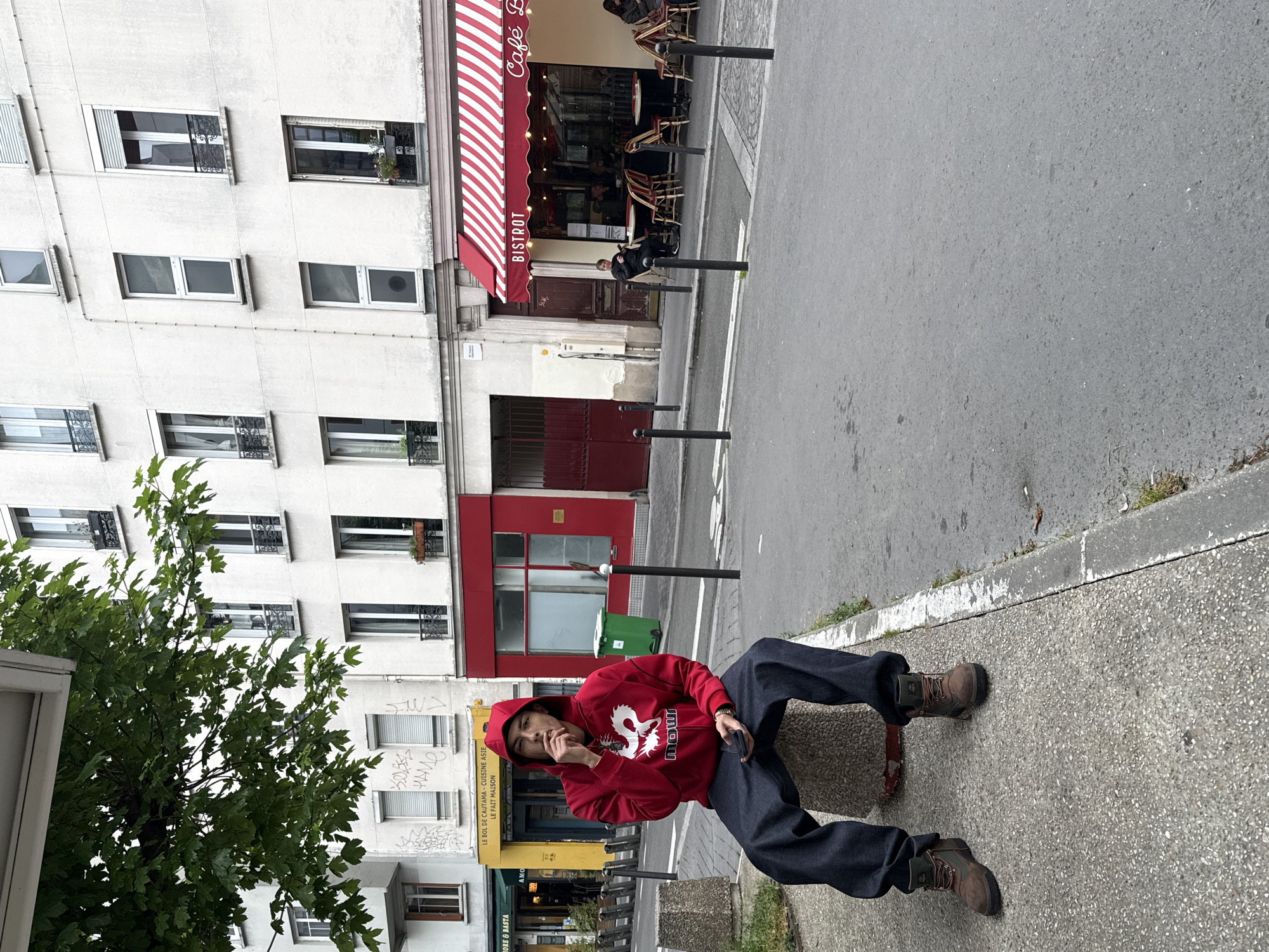 The image shows a person sitting on a concrete structure near a sidewalk in what appears to be a street in Paris, France. The person is wearing a red hooded sweatshirt, dark blue pants, and brown boots. They are holding something in their hand near their mouth.
In the background are several buildings, one of which has a red and white striped awning with the word "BISTROT" printed on it. There is a "Cafe B" sign above the entrance. Other storefronts are visible, including one with yellow signage reading "LE BOL DE CAUTAMA - CUISINE ASIE" and "LE FAIT MAISON."
The street is paved, and there are metal poles lining the sidewalk. A green trash receptacle is also present near a building with a red facade. A leafy tree is situated to the left of the frame. The sky is overcast.