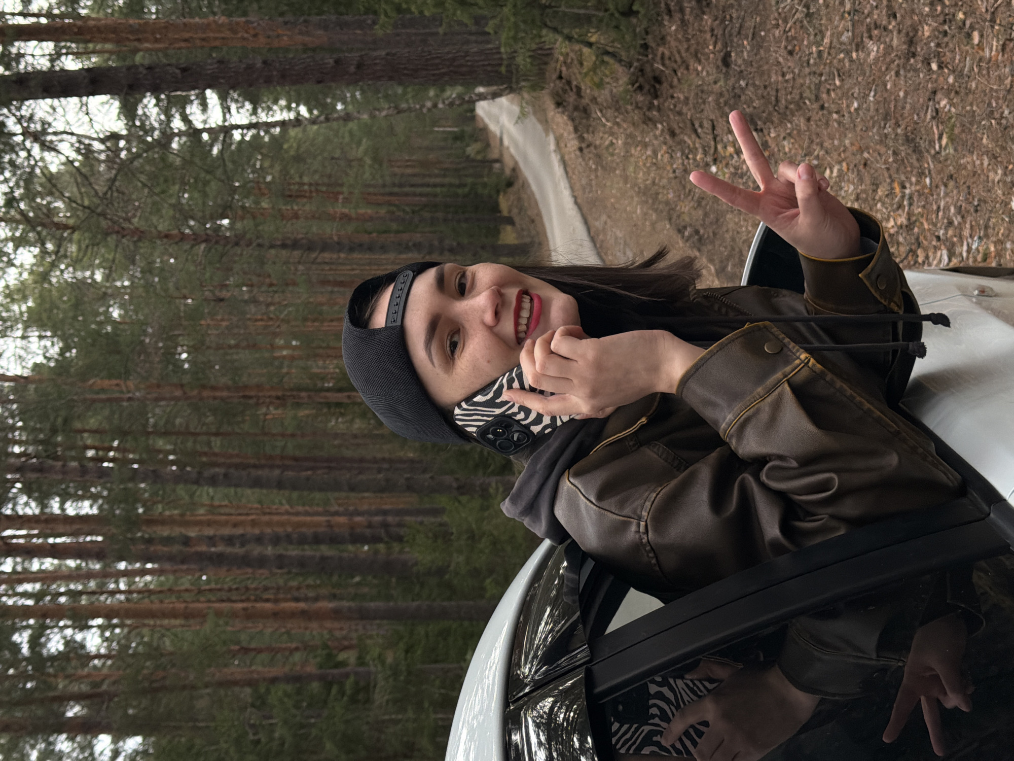 A young woman is captured leaning out of the passenger side window of a white car, smiling broadly. She has dark hair, dark eyes, and is wearing red lipstick. On her head, she wears a black baseball cap backward, and she is dressed in a brown leather jacket over a black hoodie or sweatshirt. In her right hand, she holds a smartphone with a distinctive black and white zebra print case near her ear or face, while her left hand is raised, making a peace sign. Her expression is cheerful and happy.
The setting is a dense pine forest, characterized by tall, slender trees with reddish-brown trunks and green foliage, indicating a natural environment. A narrow, unpaved dirt or gravel road winds through the forest in the background. The lighting suggests it is daytime, possibly overcast, creating soft illumination. The scene conveys a relaxed, candid moment. This image is set in the vicinity of Chaikovsky, Russia. No discernible text is visible on the woman's attire or the phone case, apart from the zebra pattern.