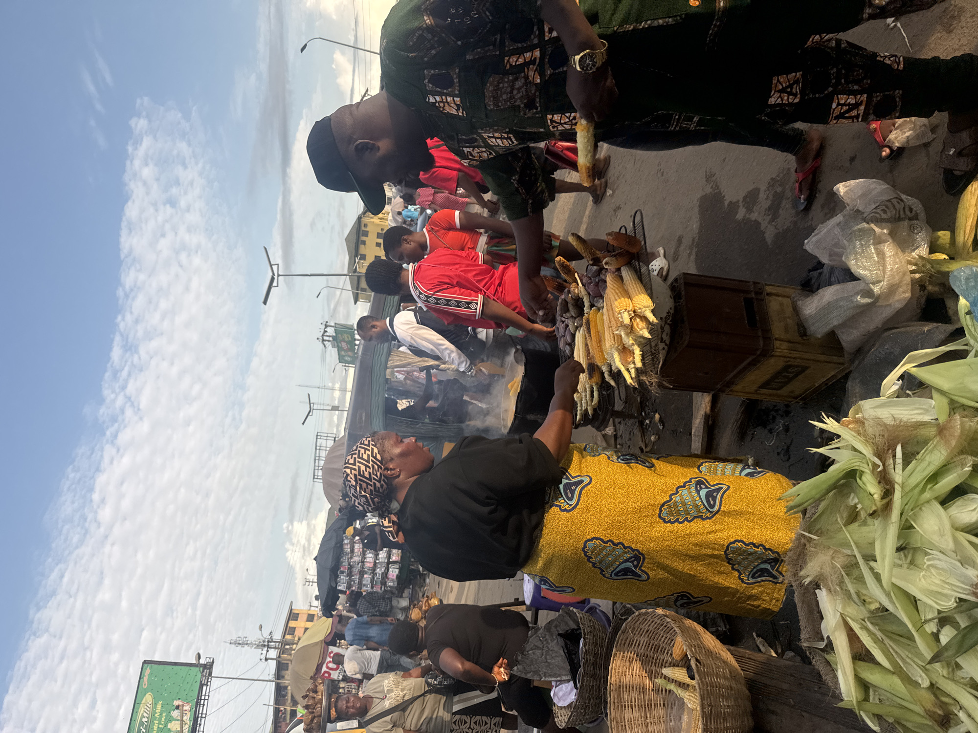 The image depicts a bustling street scene in Onitsha, Nigeria, likely in a marketplace. In the foreground, a woman in a bright yellow, patterned skirt and a black top is tending to a grill of corn, with smoke rising from the cooking. Nearby, a man in a dark green, patterned shirt and black pants is selecting a piece of corn. In the background, various people are visible, some in stalls, and a tricycle-like vehicle is parked. Stalls with umbrellas and goods for sale are set up along the street. Piles of corn husks are also visible in the foreground. The sky is partly cloudy.
