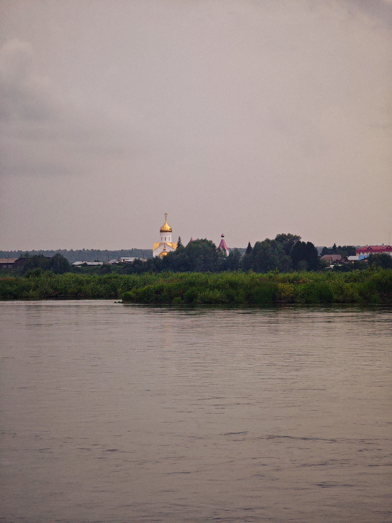 The image shows a serene, overcast scene in Kazachinskoe, Russia, likely during the evening or early morning. A calm river occupies the foreground, reflecting the muted light of the sky. The middle ground features a lush, green treeline that separates the water from a village. Prominently visible within the village is a white church with a golden dome, characteristic of Russian Orthodox architecture. To the left of the church, a smaller structure with a pink, conical roof is also present. Several residential buildings with red roofs are visible further to the right, indicating a populated area. The sky is a soft gray, with subtle variations in tone suggesting clouds. No people or immediate actions are depicted; the image captures a tranquil, static view of the landscape and settlement.