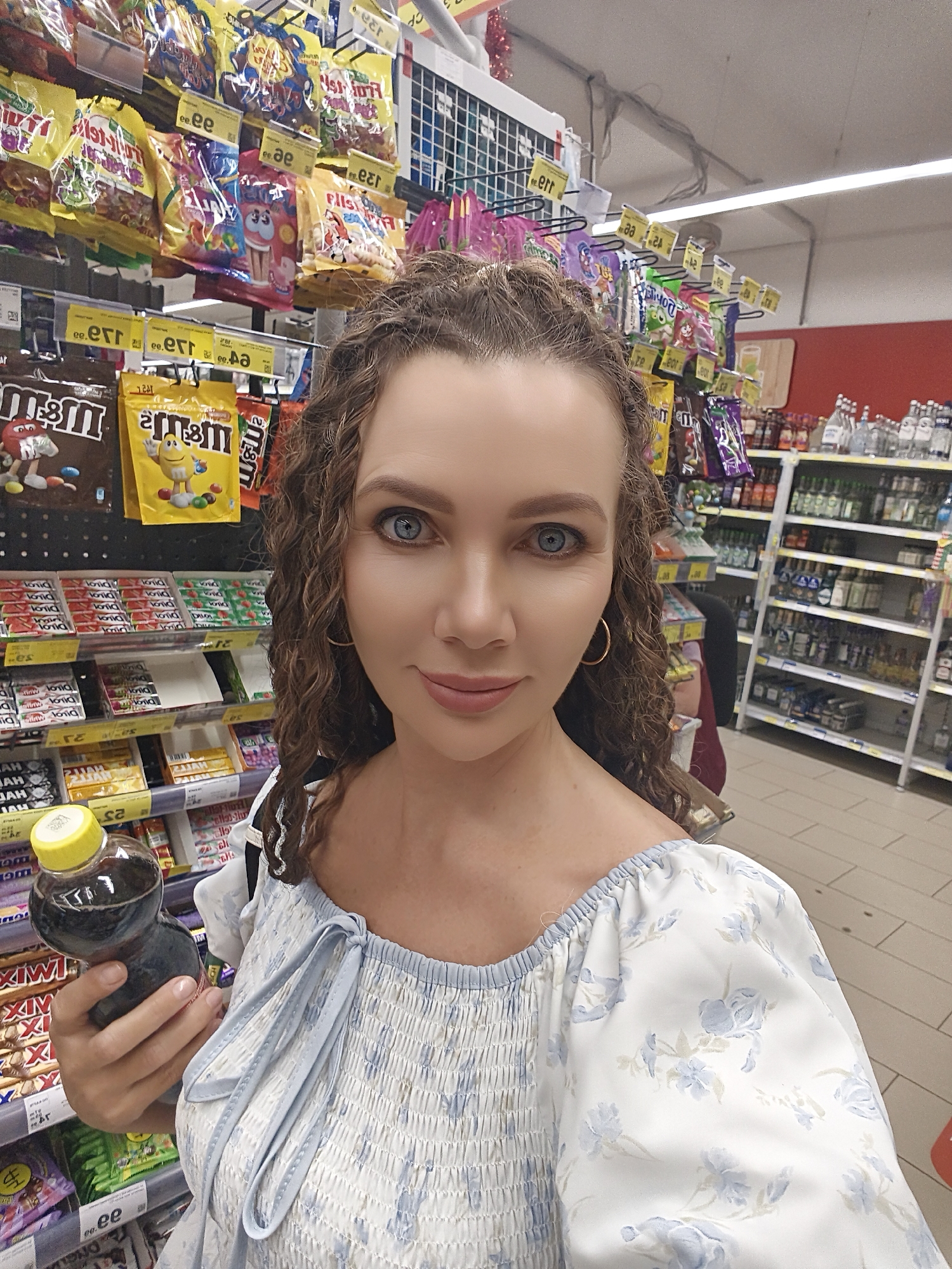 A woman with curly brown hair and blue eyes takes a selfie in a grocery store aisle in Chaikovsky, Russia. She is wearing a white and blue floral off-the-shoulder top and holding a bottle of dark liquid with a yellow cap. The shelves behind her are stocked with various candies, snacks, and bottled beverages. The aisle appears to be well-lit, and other shoppers are visible in the background.