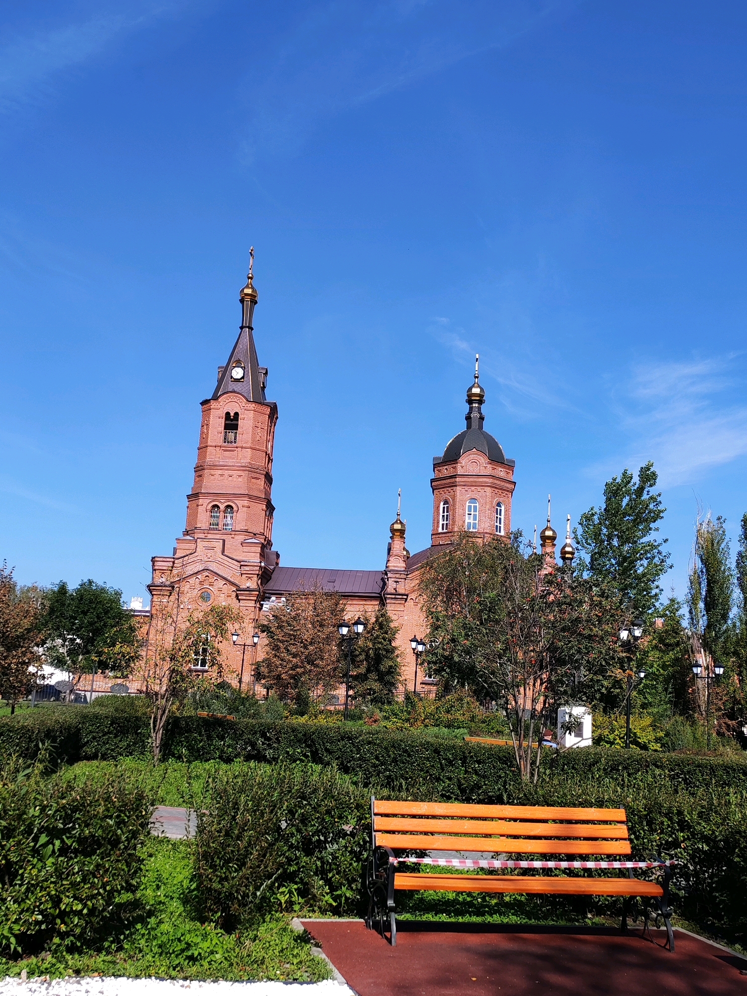 An exterior, eye-level, medium shot captures a brick church with a tall, pointed bell tower and multiple domes under a clear blue sky. In the foreground, a closed-off orange park bench sits on a dark red paved surface, with a red and white striped caution tape wrapped around its slats. Behind the bench is a manicured hedge and a lush green lawn. Several trees with autumn foliage are scattered throughout the scene, with lampposts visible in the background. The church features arched windows, decorative brickwork, and golden crosses atop its domes. The overall scene depicts a serene, somewhat deserted public space with a prominent religious structure.