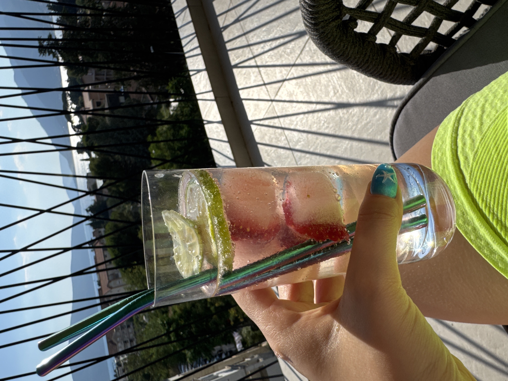 A person's hand, with teal nail polish and a starfish design on one fingernail, holds a tall glass filled with a pink beverage, ice cubes, and lime slices. Two iridescent metal straws extend from the glass. The background shows a balcony with a railing, casting shadows on the tiled floor, and a view of a body of water with distant hills and buildings under a bright blue sky. The person is wearing a neon green garment.