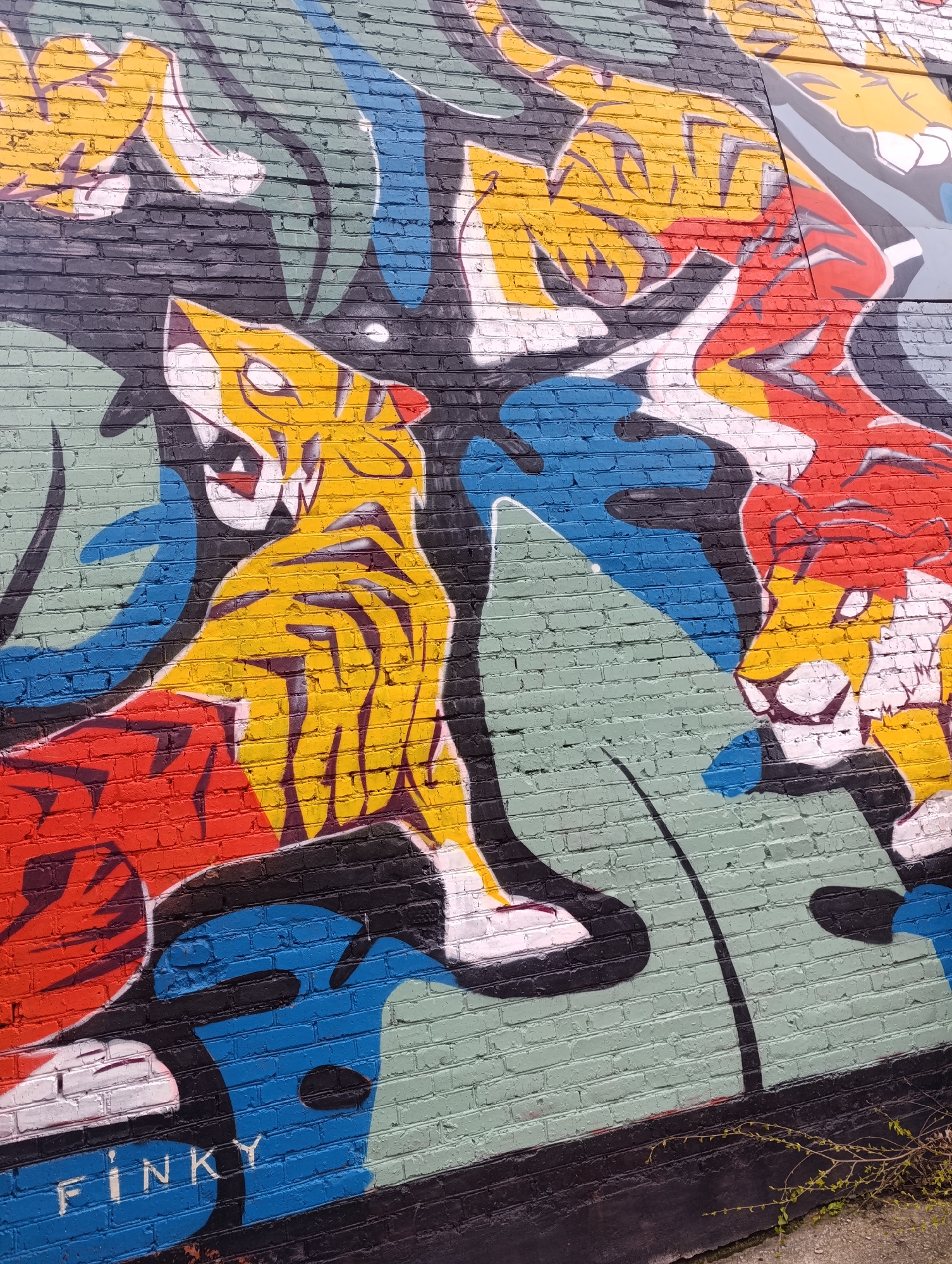 The image shows a close-up of a colorful mural painted on a brick wall. The mural depicts stylized tigers in various colors, including yellow, red, white, and black. The tigers are surrounded by abstract shapes and lines in blue, green, and gray, possibly representing foliage. The signature "FINKY" is visible in blue paint at the bottom left of the wall. The overall impression is of a vibrant and energetic street art piece.