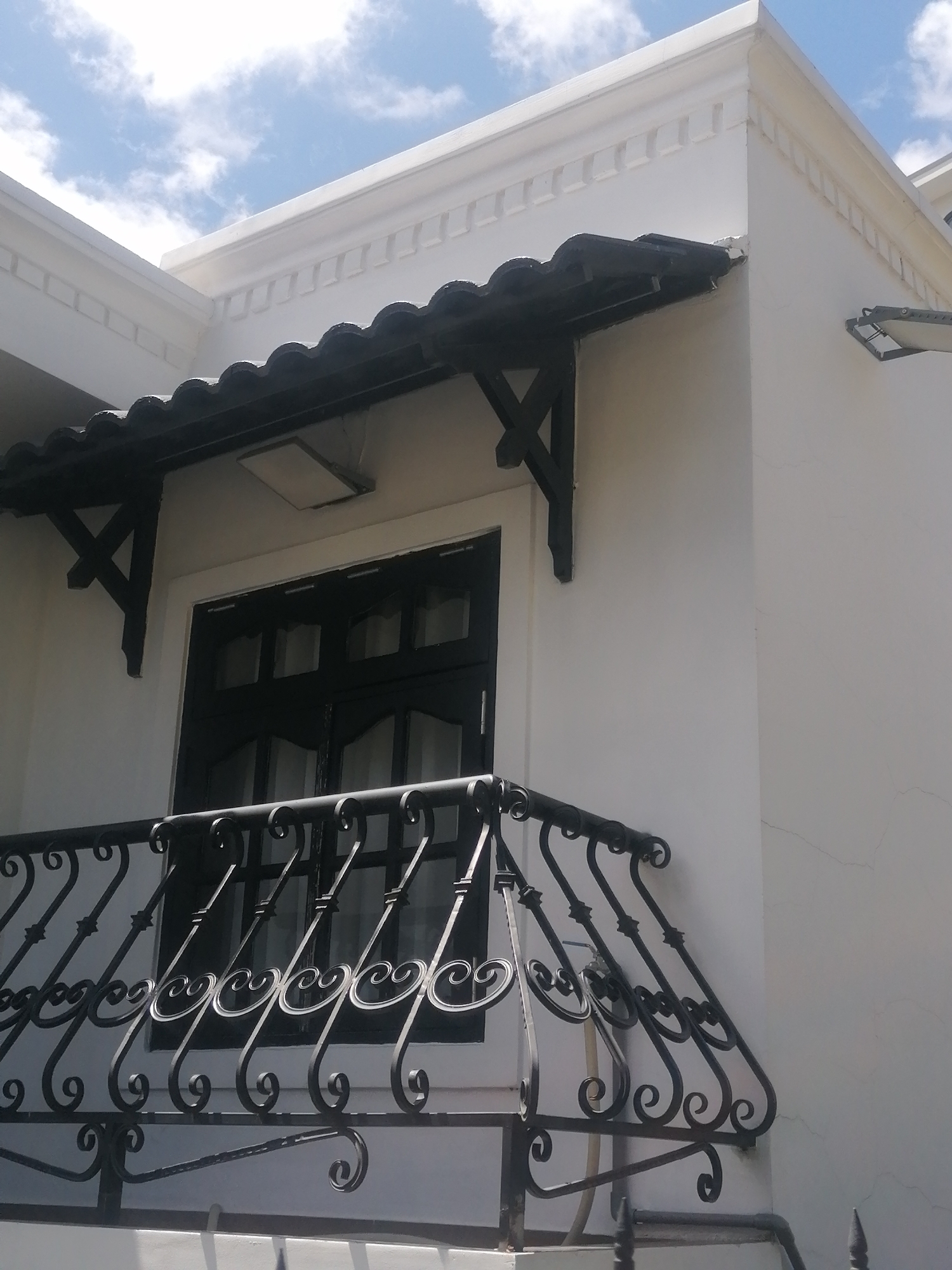 This is a close-up, eye-level, exterior shot of a white building in Da Lat, Vietnam, under a bright blue sky with scattered white clouds. The building features a black tiled awning supported by dark decorative brackets above a black-paned window. Below the window, a black wrought-iron balcony railing with ornate scrollwork is visible. The architectural style suggests a residence or a small commercial establishment. The scene is clean and well-maintained, with no visible people or signs of activity. The lighting indicates it is daytime, likely mid-morning or afternoon, given the angle of the sun. The overall impression is one of a quiet, picturesque urban facade.