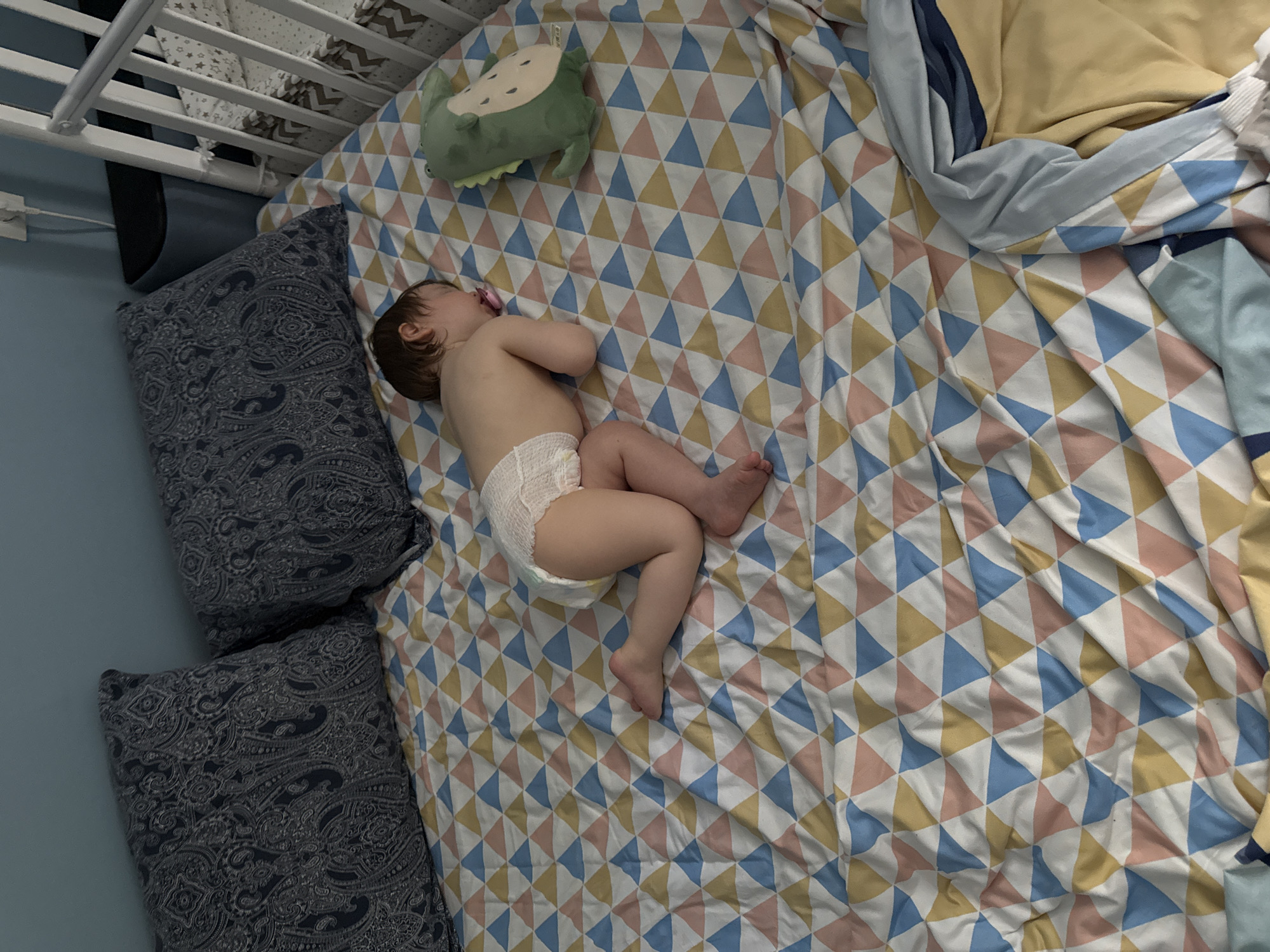 A baby is sleeping in a crib. The baby is wearing a diaper and has a pacifier in its mouth. A green toy is next to the baby. The crib is covered with a triangular patterned blanket. Pillows are near the crib. The location is not discernible from the image.
