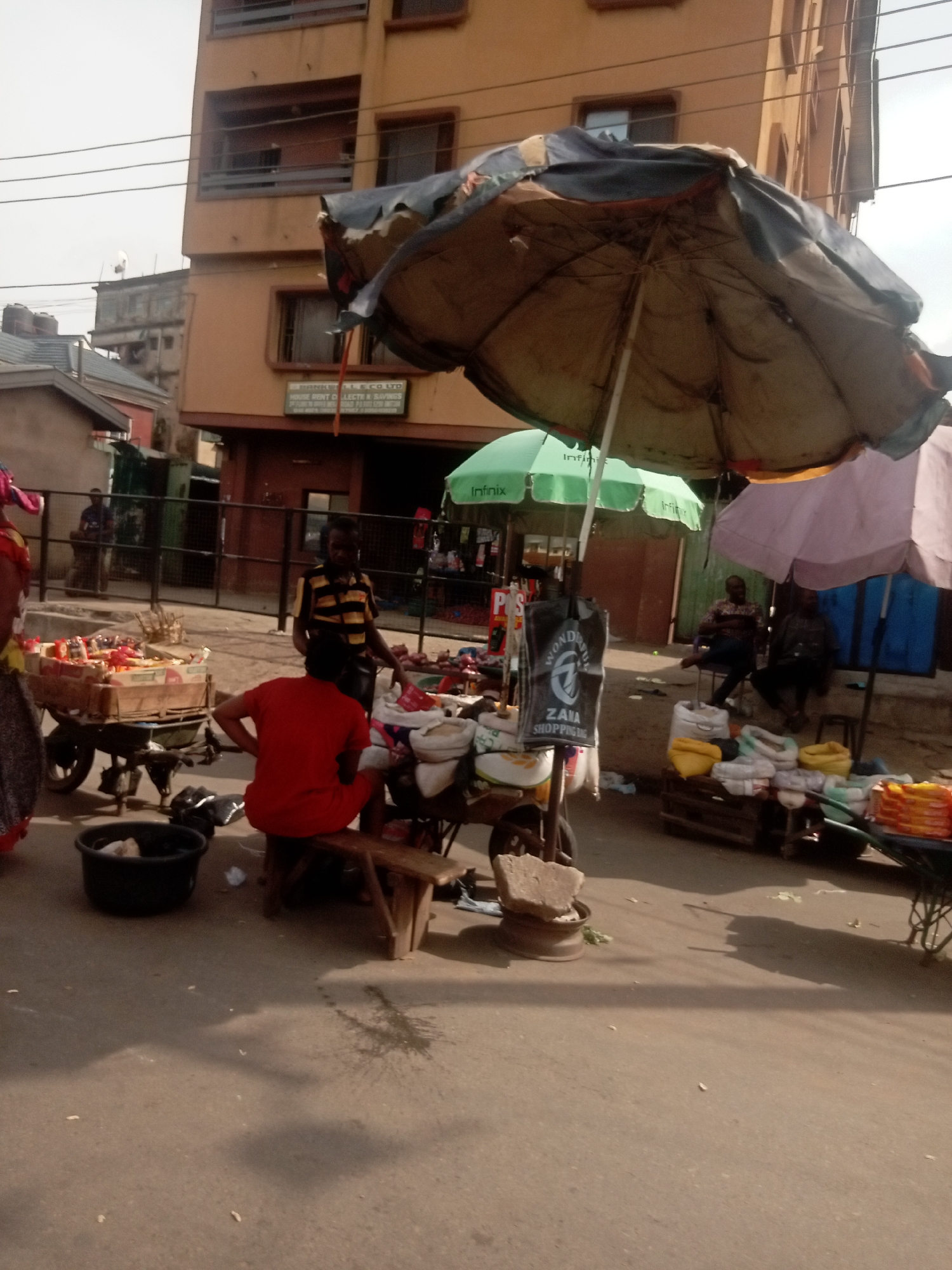 The image depicts a street scene in Onitsha, Nigeria. Multiple market stalls are visible, each sheltered by umbrellas. One stall features a man in a striped shirt, standing behind a cart laden with goods. Another stall displays sacks of items, possibly grains or produce. A person in a red shirt sits on a small bench near a black bucket. In the background, two men are seated, while a building is visible above the market stalls. The overall environment suggests a bustling commercial area.