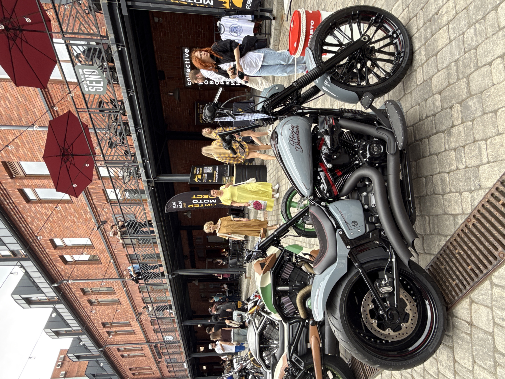 This is a full-shot, eye-level, outdoor photograph of a motorcycle event likely held in Saint Petersburg, Russia, given the Cyrillic text visible on banners and signs. In the foreground, a custom grey Harley-Davidson motorcycle is prominently displayed. It features a large rear tire, black exhaust pipes, a black and red seat, and a "Harley Davidson" logo on the fuel tank.
In the mid-ground, several other motorcycles are parked, including one with a camouflage green and brown paint scheme. A group of people are milling around, some looking at the motorcycles, others conversing. A woman in a yellow dress and another in a brown dress are visible, along with men in casual attire. Behind the motorcycles, a brick building with multiple levels forms the backdrop. Red umbrellas are visible on an upper balcony, and signs in Russian are displayed on banners. One banner, with Cyrillic text and a date "31.07 02.08 2026," suggests a specific event. Another sign reads "collective."
The environment appears to be a paved outdoor area with brick buildings characteristic of an urban setting. The lighting suggests it is daytime, possibly overcast. The overall scene depicts a gathering centered around custom motorcycles, with an urban, possibly historic, district as the location.
