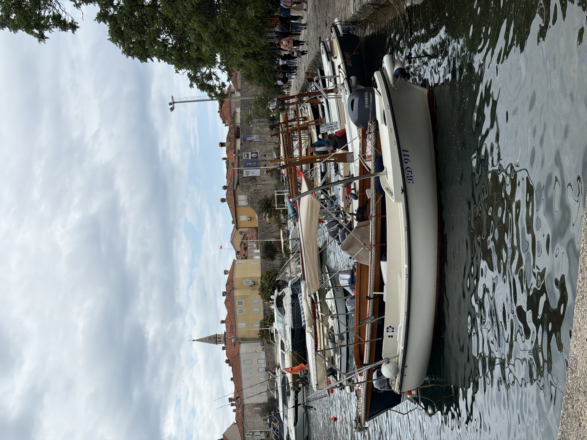 A horizontal photograph shows a row of docked boats and a waterfront walkway with buildings in Budva, Montenegro.
The boats vary in size and style, and are mostly white with some visible engine parts, rigging, and identification markings like "BD 911". They are docked along a concrete pier or walkway, with visible reflections in the water.
Behind the boats, a walkway is lined with people, some of whom are observing the boats or the scenery. The buildings behind the walkway are a mix of old stone structures and more modern constructions with yellow facades and red-tiled roofs. A church steeple is visible among the buildings.
The sky is overcast, with some glimpses of blue. A tree is visible at the top, its branches partially obscuring the sky.