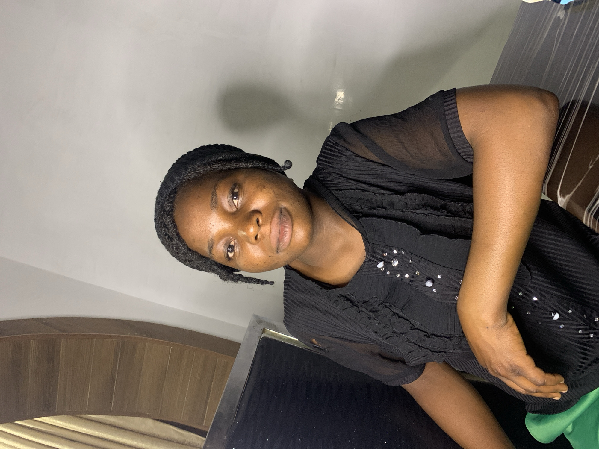 The image shows a woman with dark skin, braided dark hair, and brown eyes, wearing a black sheer blouse with ruffled detailing and bejeweled buttons. She is seated at a dark, glossy table, with her left arm resting on it. A green garment is visible on her left side. The background features a light-colored wall and a wooden curved panel with vertical slats. The scene appears to be indoors, possibly in a residential or commercial setting in Jos, Nigeria.
