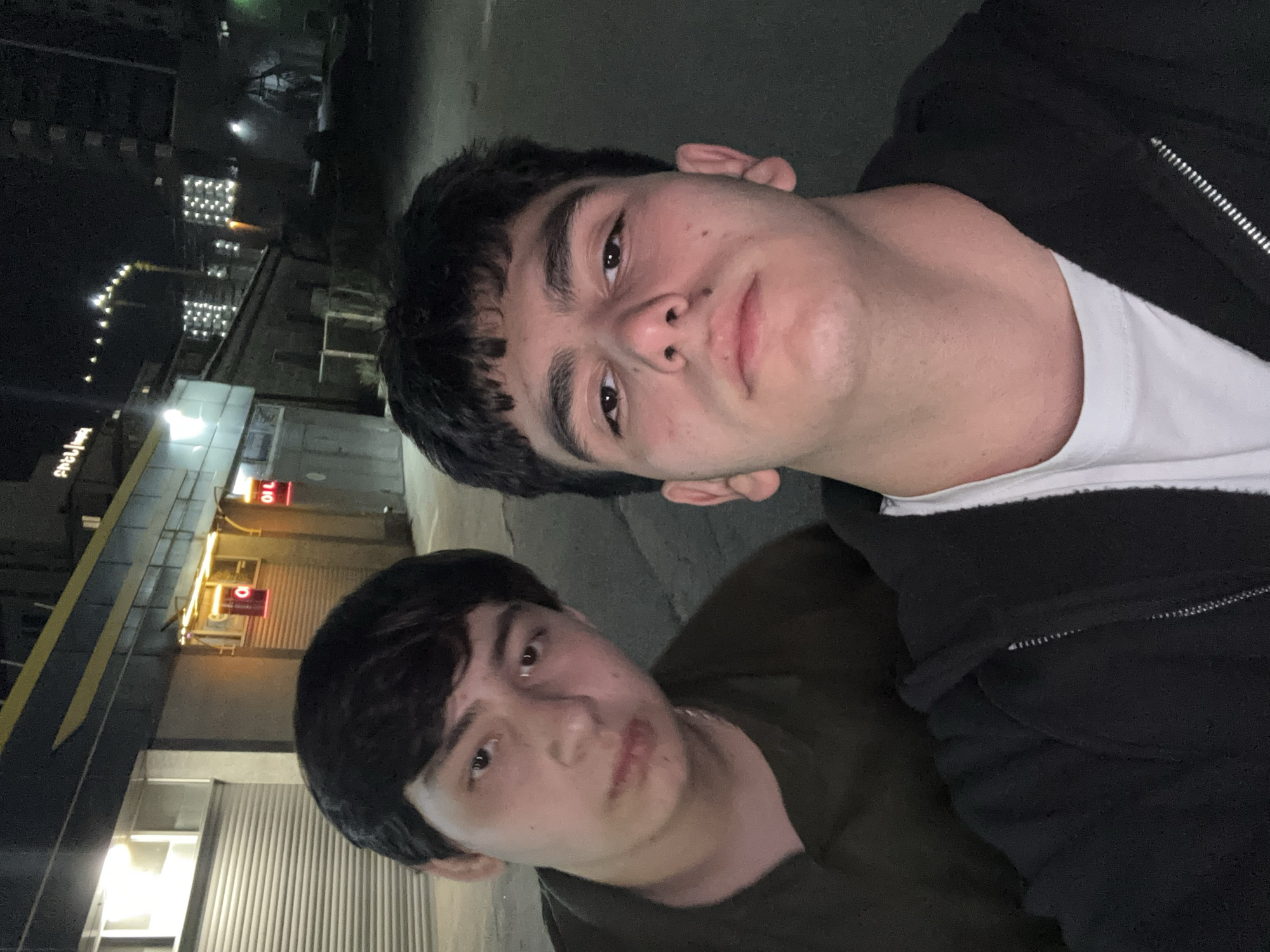Two young men are posing for a selfie at night in Yerevan, Armenia. The man in the foreground is wearing a dark green shirt. The man behind him is wearing a white t-shirt and a black jacket. A building with a yellow awning and a red "OIL" sign is visible in the background. Streetlights and illuminated buildings can be seen in the distance.