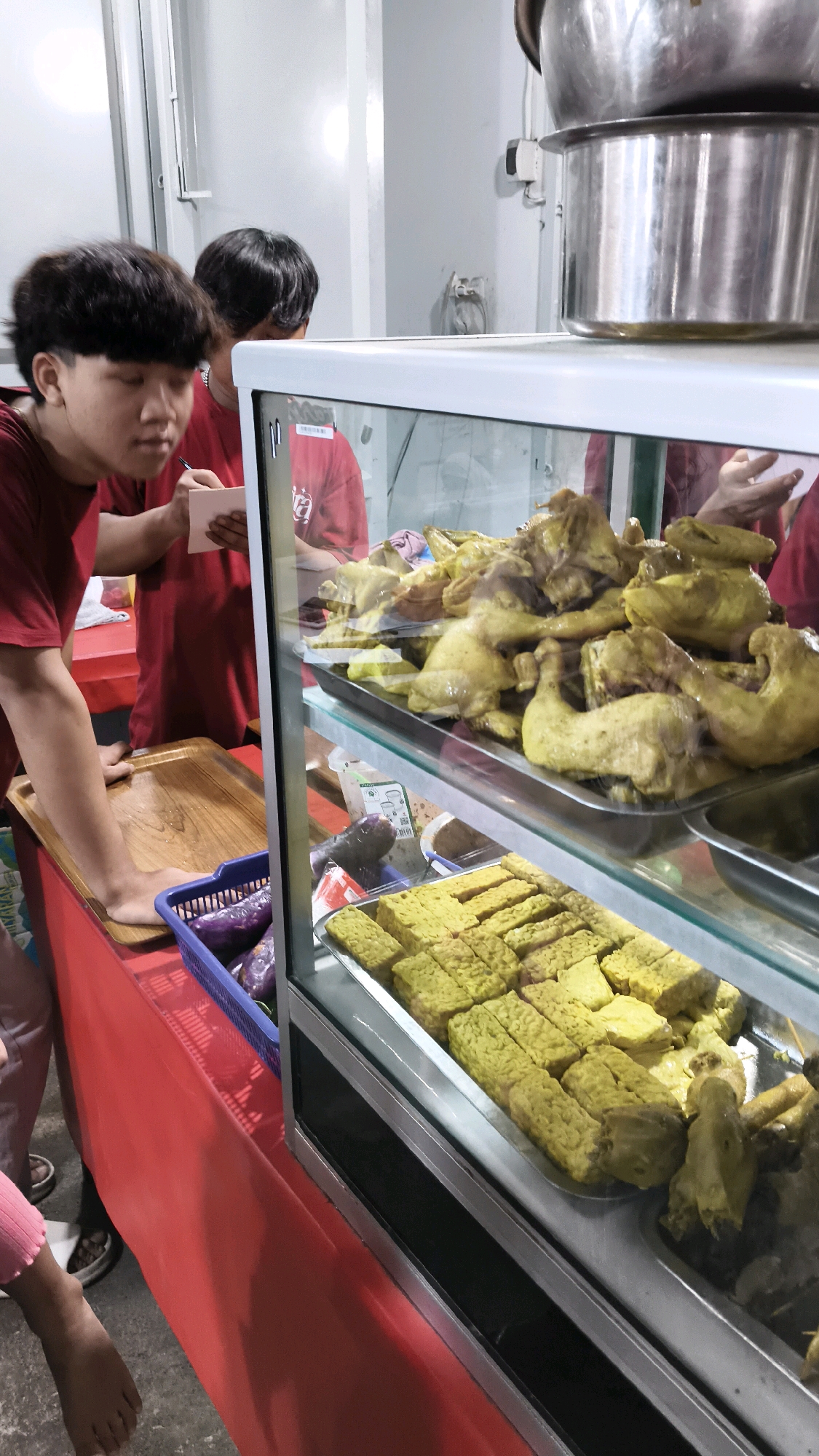 In Karangsatria, Indonesia, a food stall is depicted. The stall features a glass display case filled with food. On the top shelf of the case, there are trays of cooked chicken pieces that appear to be boiled or steamed, with a light yellow hue. Below the chicken, on the second shelf, are rectangular pieces of fried tempeh, also yellowish-green. To the left of the display case, a person wearing a red t-shirt is reaching for a wooden tray. Another person in a red t-shirt is standing behind them, holding a small white notepad and a pen, possibly taking an order or recording sales. A blue plastic basket containing purple vegetables, possibly eggplants, is visible on the counter to the left of the display. The stall is set up with a red tablecloth covering the counter area. In the background, a wall and part of a kitchen or service area are visible, including large metal pots stacked on top of each other. The lighting suggests an indoor setting, possibly a market or a covered food court.