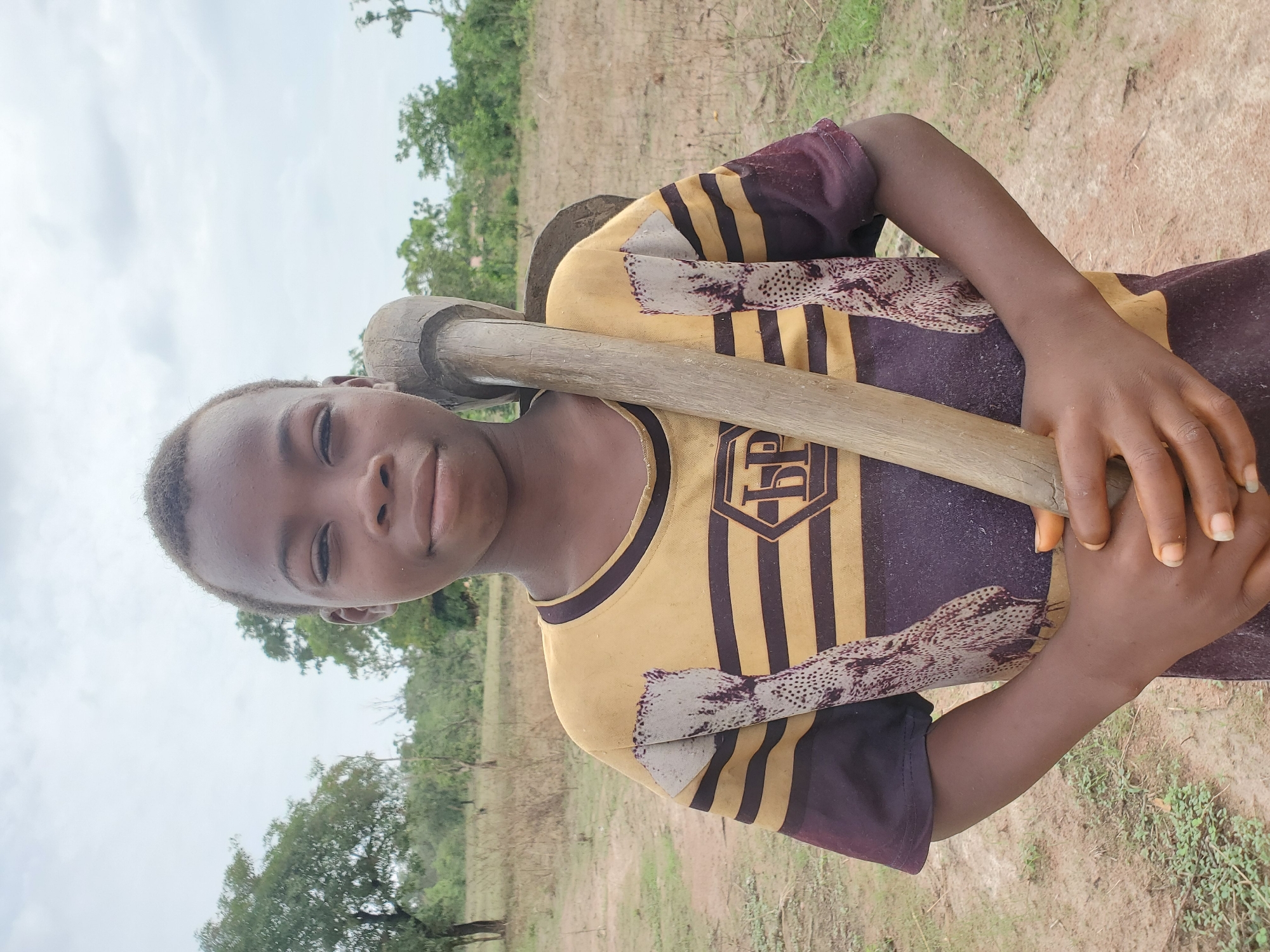 A portrait-style image features a young person, likely a child or young adolescent, with dark skin and short, dark hair, looking directly at the camera. They are positioned outdoors in what appears to be a rural or semi-rural setting in Mararaba, Nigeria.
The subject is wearing a two-toned t-shirt, primarily yellow with horizontal dark-colored (possibly maroon or dark brown) stripes on the sleeves and body. A faded design or logo, which includes a stylized letter (possibly 'P' or 'PD') within a shield shape, is visible on the chest area of the yellow fabric.
They are holding a large, rustic wooden farming tool, resembling a hoe or mattock, over their right shoulder. The long wooden handle extends diagonally across their body, and their hands are wrapped around it. The tool's head, also made of wood, rests behind their head.
The background consists of dry, light brown soil or dirt, indicative of an uncultivated field or ground. Scattered sparse green vegetation, including trees in the mid-distance and small plants on the ground, are visible. The sky is bright and overcast, suggesting daylight. The overall expression of the person is neutral or slightly serious. No specific activity beyond posing for the camera is depicted. The visible text on the shirt is partially obscured and faded.