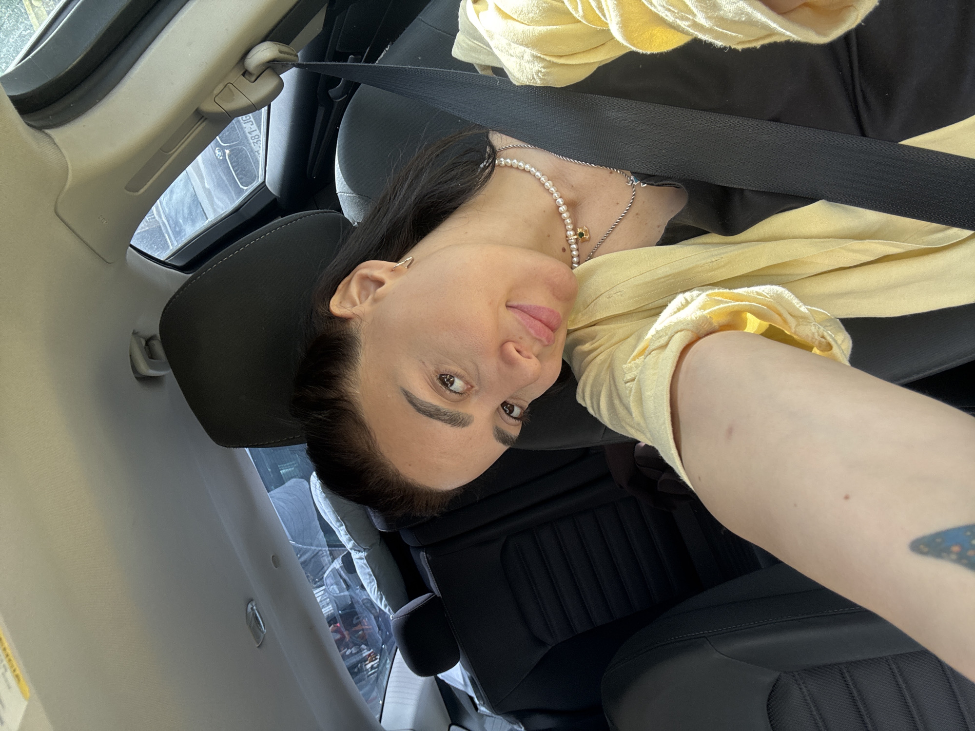 A woman is in the passenger seat of a car. She is wearing a yellow shirt, a pearl necklace, and a seatbelt. Her arm is visible in the foreground, with a tattoo on her inner forearm. The car's interior is visible, including the dashboard and seats. Through the window, a street scene is visible, possibly in Tbilisi, Georgia.