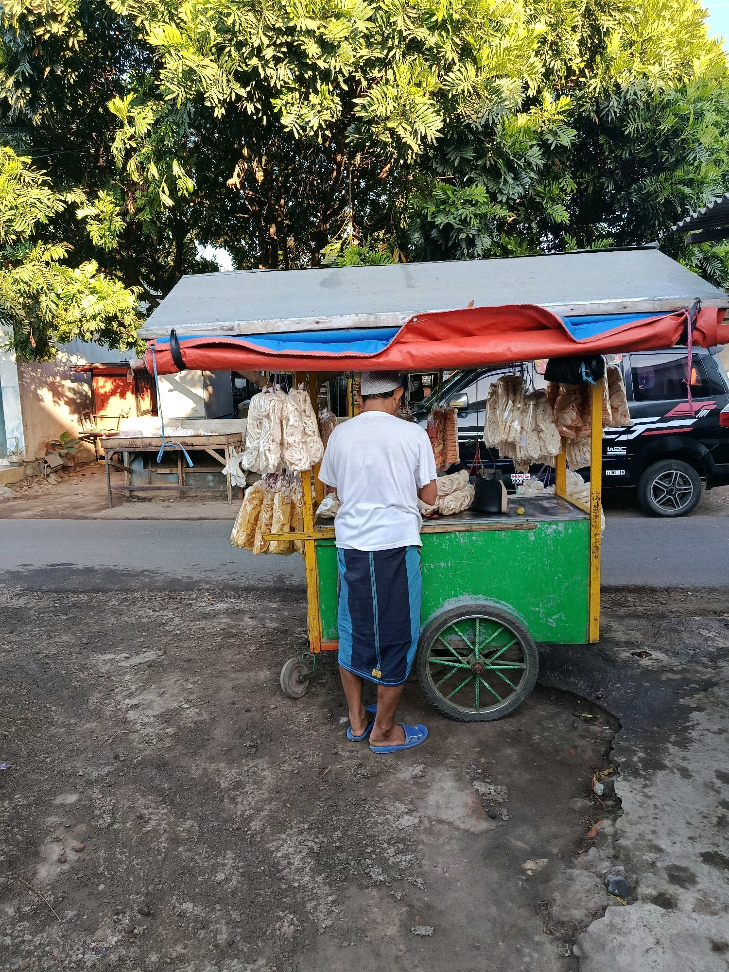 An elderly Indonesian man, wearing a white t-shirt, a skullcap, and a sarong, stands behind a green and yellow mobile food cart on a street in Bululawang, Indonesia. The cart is covered by a red and blue tarp and displays various food items hanging from racks, likely crackers or snacks. A black van is parked behind the cart. The street appears to be paved with asphalt, and the surrounding area features trees and buildings. The ground is a mixture of dirt and gravel, with some debris present. The lighting suggests it is daytime, possibly late afternoon, with the sun casting shadows.