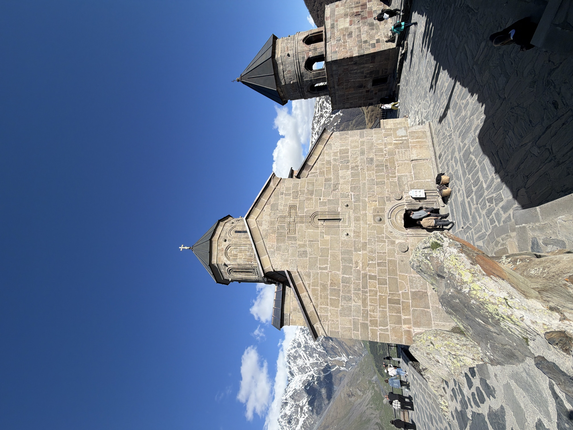 The image depicts the Gergeti Trinity Church in Stepantsminda, Georgia, under a clear blue sky with some clouds. The stone church is the primary subject, with its cross-topped structure and adjacent bell tower. The church is situated on a rocky mountain ridge with snow-capped mountains visible in the background. People can be seen both near the entrance of the church and along a lower walkway, suggesting tourist activity. The foreground includes parts of the stone ground, further emphasizing the mountainous terrain on which the church stands.