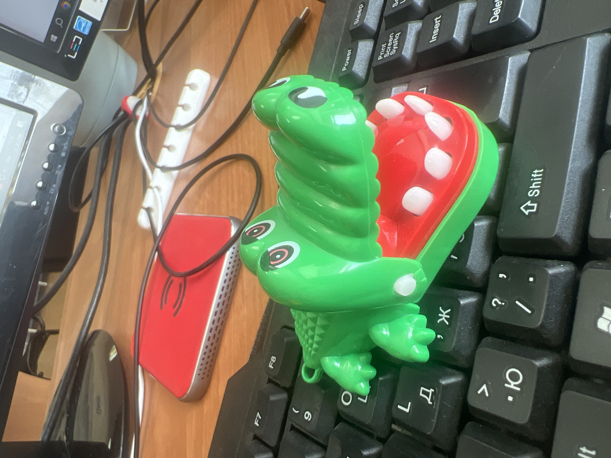 A green toy alligator with red jaws and white teeth sits on a black keyboard. The alligator's body is textured and it has large, cartoonish eyes with red and white irises. The keyboard has Cyrillic lettering on some of the keys, indicating it is likely a Russian keyboard. In the background, a red object, possibly a phone or a case, is visible, along with computer monitors and various cables. The scene appears to be an office or home desk environment. The city of Omsk, Russia, is mentioned in the prompt, but there are no specific visual cues in the image that confirm its location within Omsk.