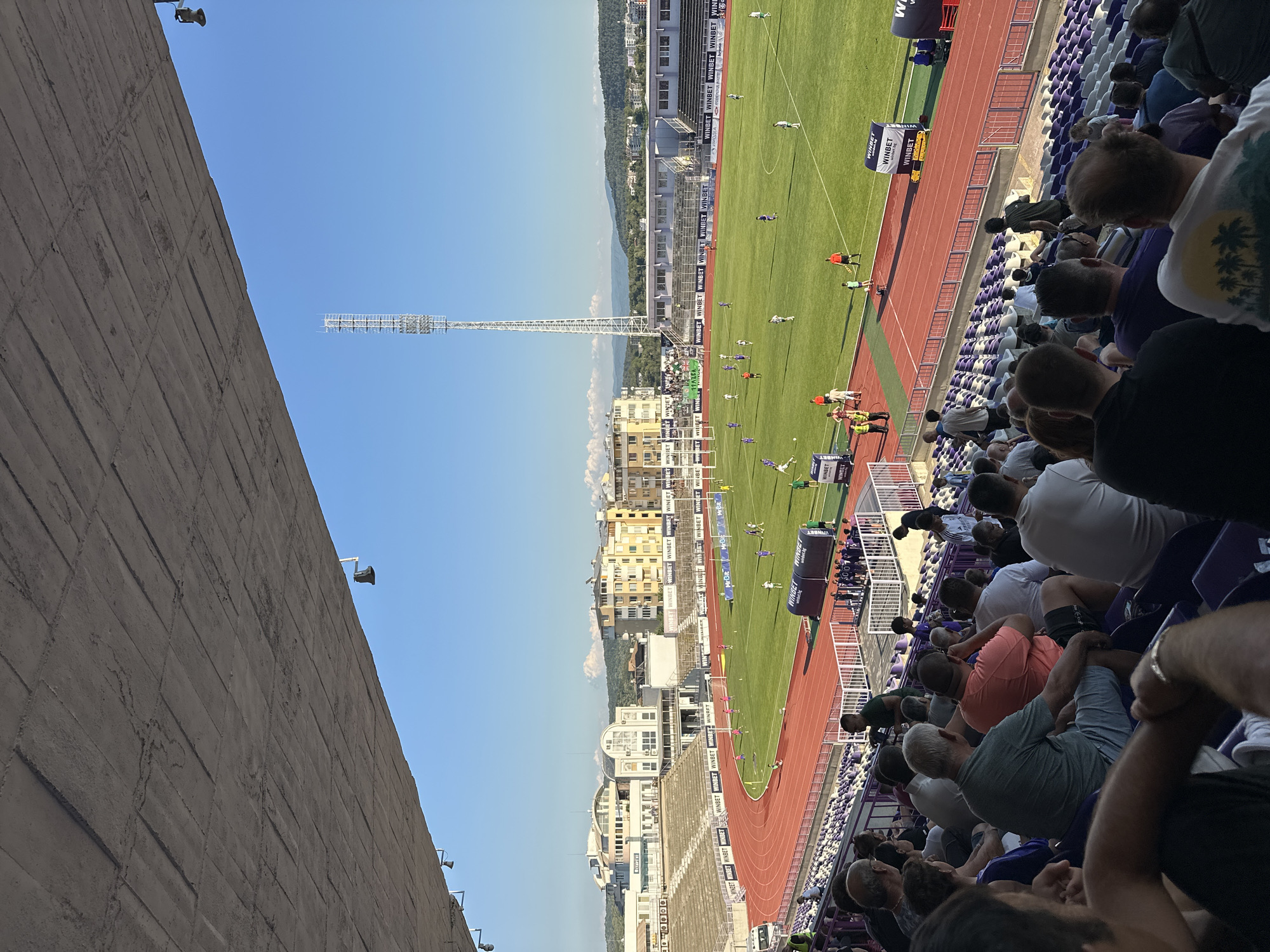This is an aerial, eye-level, full shot of a football match taking place in the stadium in the city of Veliko Tarnovo, Bulgaria. The stadium has a running track surrounding the grass field. The stands, filled with spectators, are visible on the left and in the foreground. The spectators are seated in purple and white chairs. On the field, several players in white and purple uniforms are engaged in the match. A tall broadcast tower stands in the background, with buildings and hills beyond it under a clear blue sky. The image is taken from a slightly elevated position, looking down at the field and across the stadium.