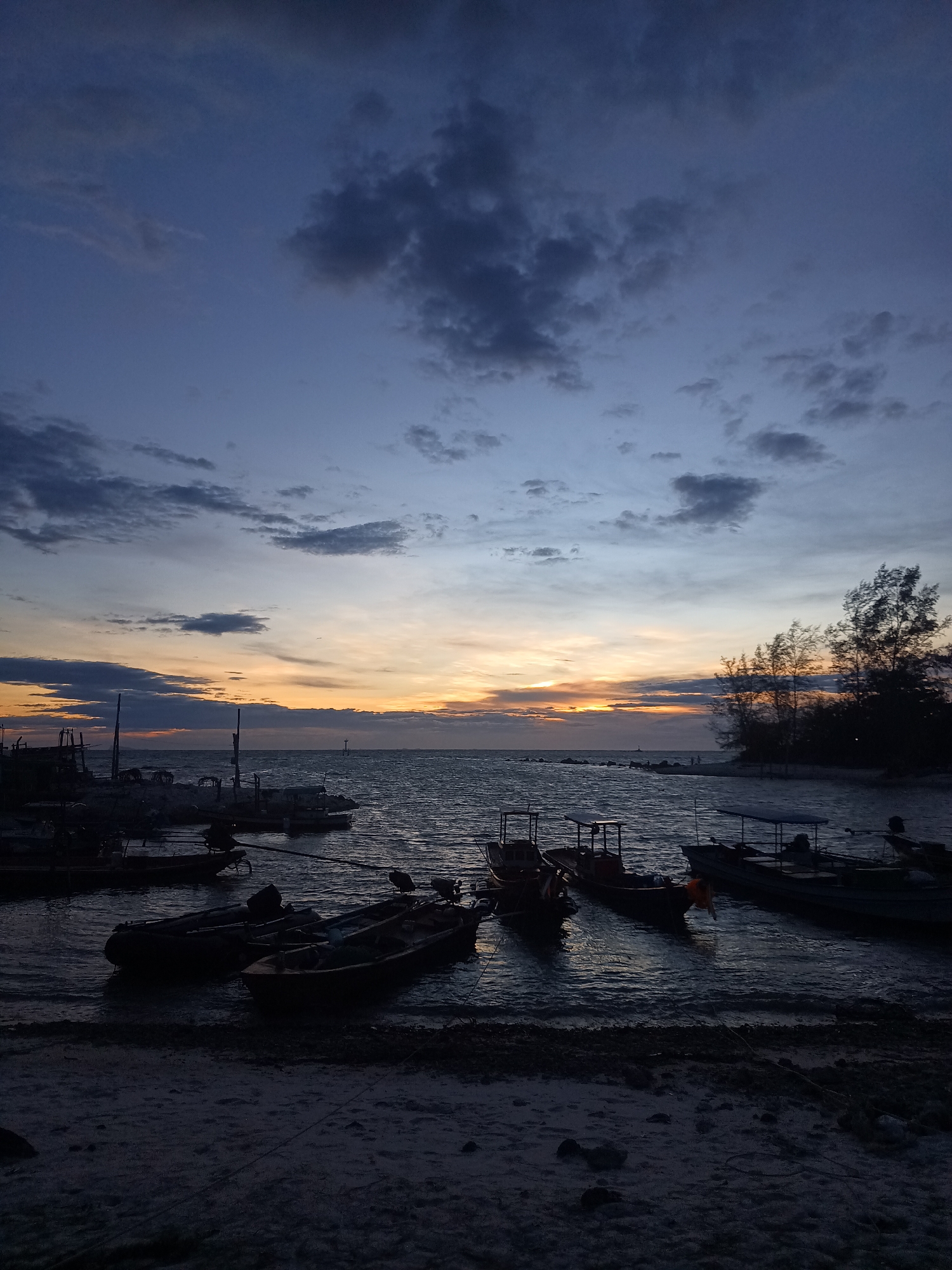 The image depicts a twilight scene at Baan Sri Thanu, Thailand. The sky shows a gradient from dark blue to orange at the horizon, with scattered clouds. Several boats are near the shore, some close to the sand, and others moored further out. Trees are visible to the right, silhouetted against the sky. The foreground consists of a sandy beach.