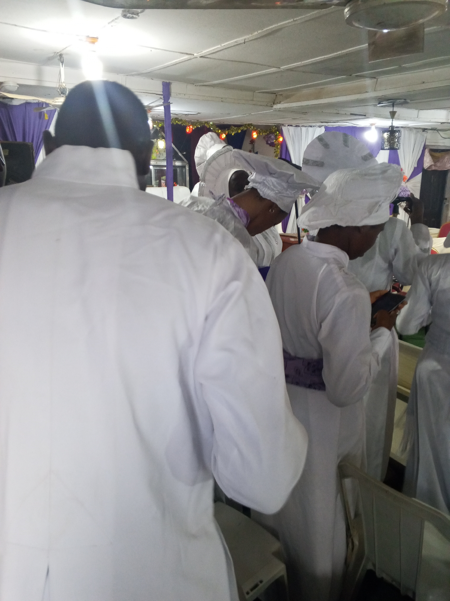 The image shows a gathering of people inside what appears to be a church or religious hall in Lokoja, Nigeria. Several individuals are wearing white robes and head coverings, which suggests they may be part of a religious order or participating in a specific ceremony. One person in the foreground is facing away from the camera, revealing the back of their white robe. Another individual is looking at a mobile phone. The environment is illuminated by artificial lighting, with some decorations visible in the background, including draped fabrics in white and purple. The overall atmosphere seems to be one of religious observation or gathering.