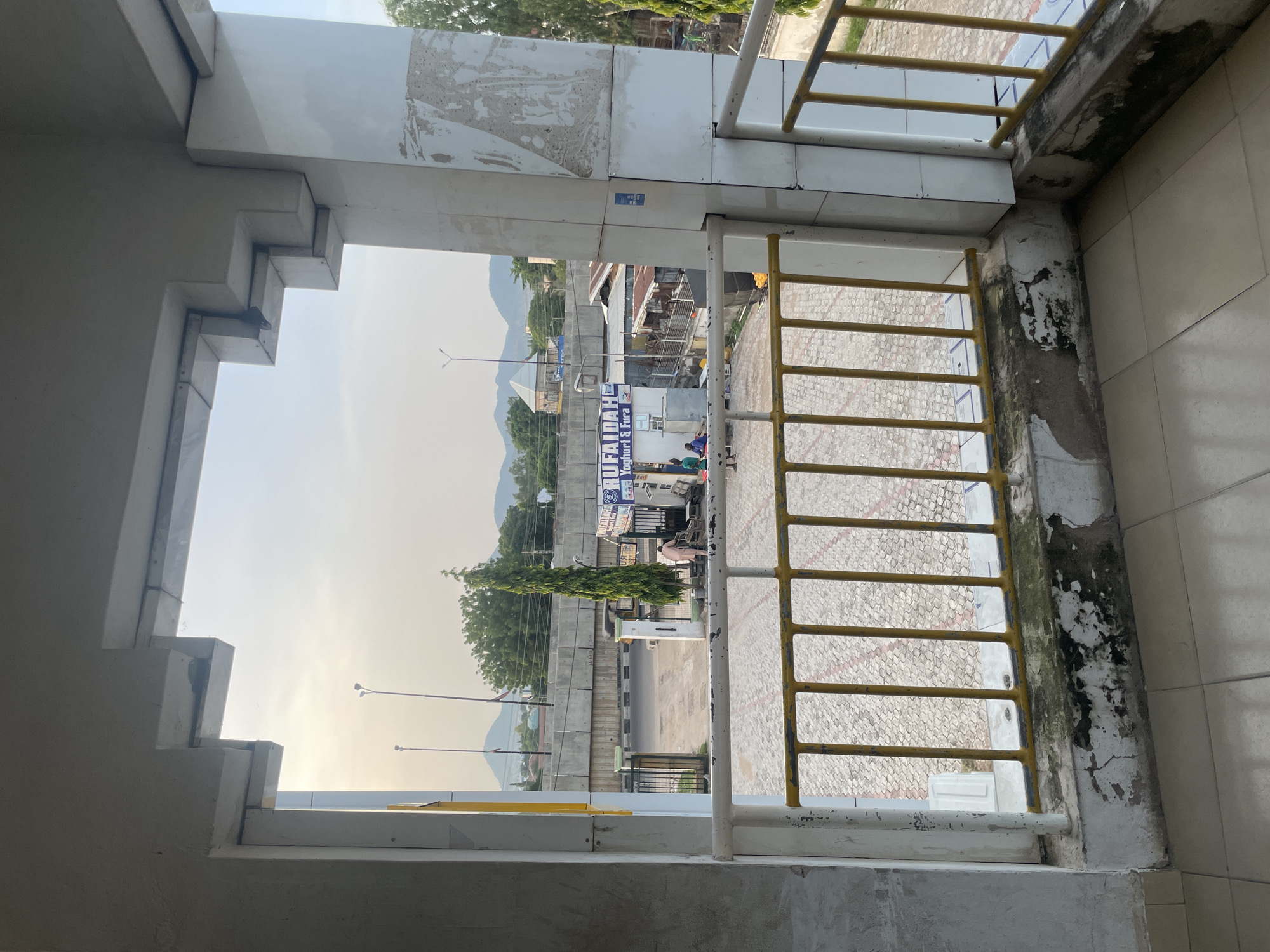 An outdoor view from a covered walkway in Jalingo, Nigeria, reveals a street scene under a hazy sky with distant mountains. A yellow metal railing with white vertical supports frames the foreground, suggesting a balcony or elevated walkway. Beyond the railing, a paved area with a patterned brick design is visible. Several people are present in the middle ground, near an open gate leading to a building complex. One person in light-colored clothing is walking away from the camera toward the gate, while others are gathered near the entrance. A tall, slender tree stands near the gate, and a sign with text, including "RUFAIDAH Yogurt & Fura," is mounted on a building. A road passes behind the building complex, with a concrete barrier and a pedestrian bridge overhead. Streetlights are visible, and the overall atmosphere suggests an urban environment in daylight. The architectural elements of the walkway, particularly the stepped concrete trim on the overhead structure, are prominent. The lower section of the walkway wall shows signs of wear and peeling paint.