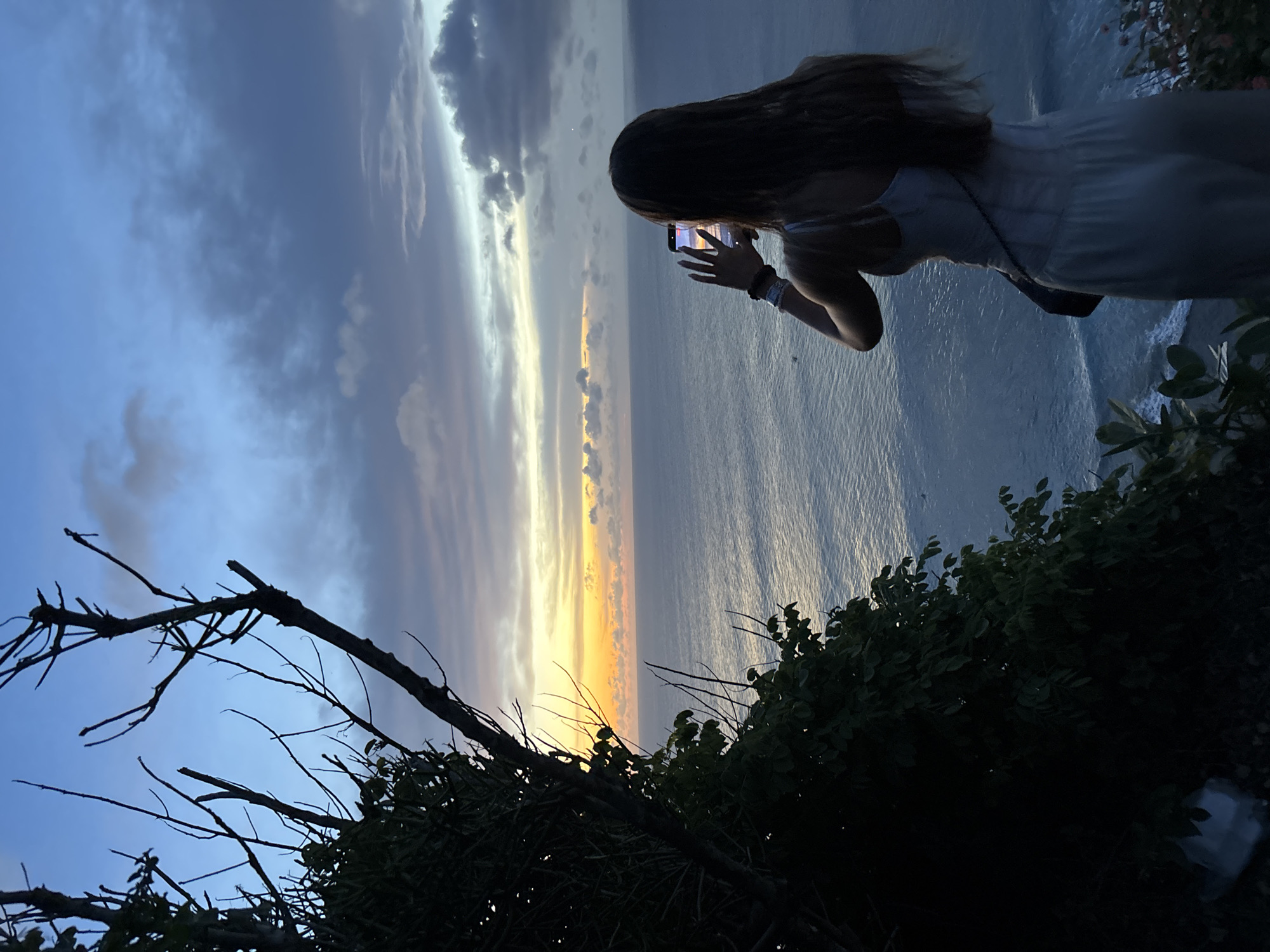 A sideways medium shot depicts a person taking a photo with their smartphone. The person is standing near a cliff, looking out over the ocean. The sky is a mix of dark blue and grey clouds with a strip of orange and yellow on the horizon, indicating either sunset or sunrise. The person has long brown hair, a white dress, and is wearing a bracelet. The foliage is in the foreground, with some branches and green leaves. The location is Uluwatu, Indonesia, as evidenced by the ocean view and vegetation commonly found in that area.