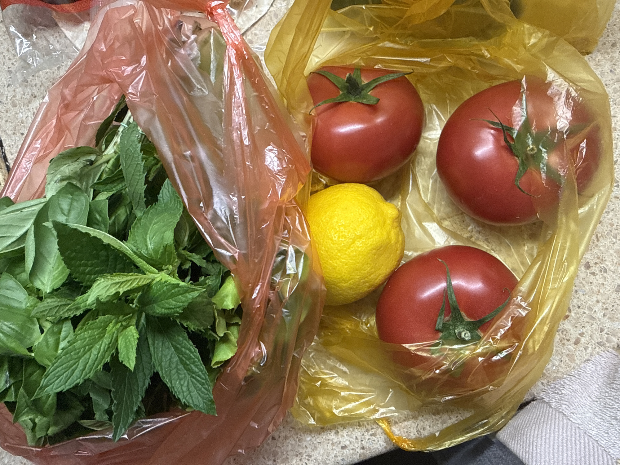 A pile of fresh herbs, including mint and basil, sits in a red plastic bag on a speckled countertop. Next to it, in a yellow plastic bag, are three ripe red tomatoes and one bright yellow lemon. The scene appears to be an indoor setting, likely a kitchen, given the countertop and the presence of grocery items. No people or actions are depicted. The environment is simple and focused on the produce.